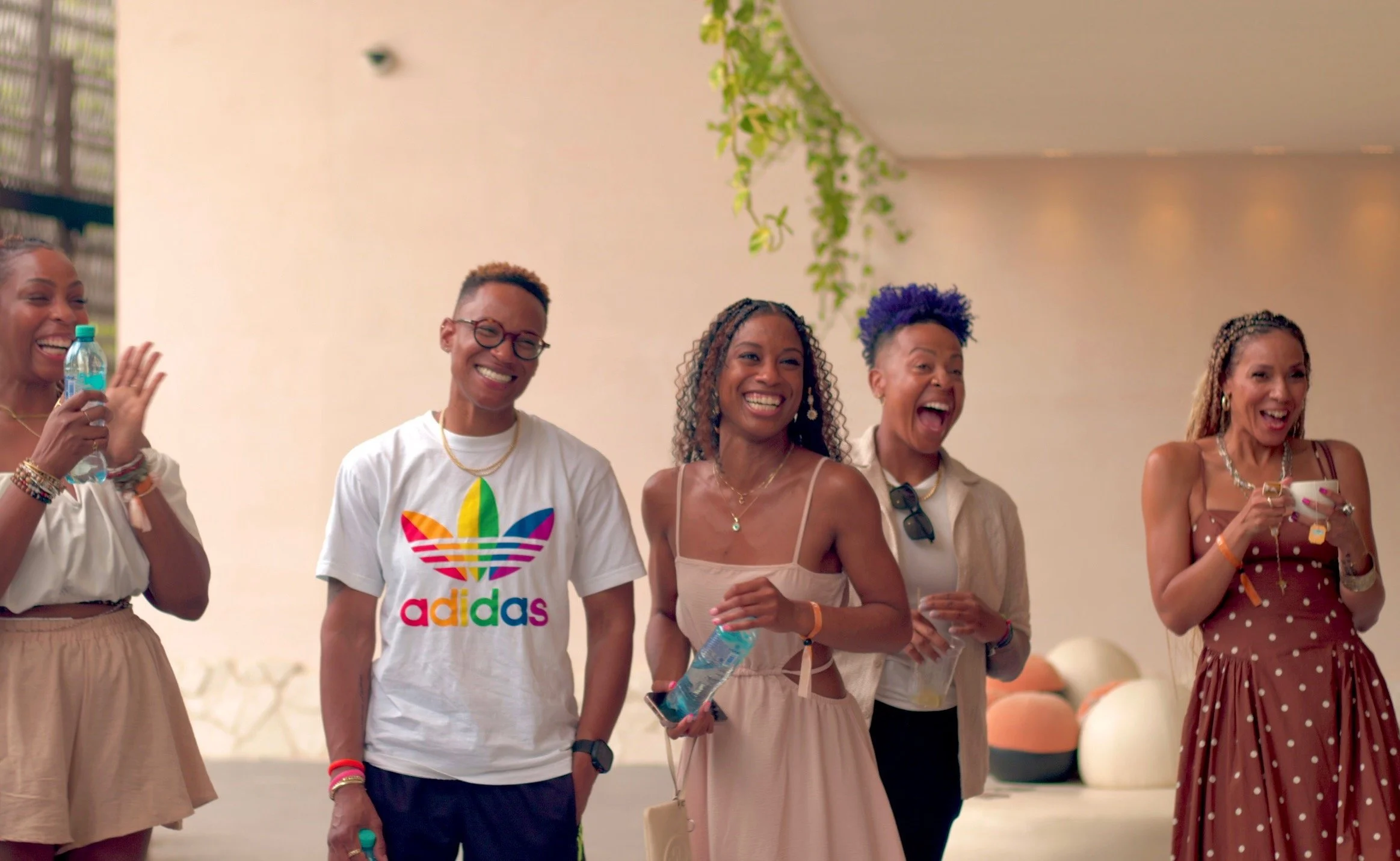 Group of five women laughing and enjoying a social gathering indoors, with a neutral beige background and some hanging green plants.
