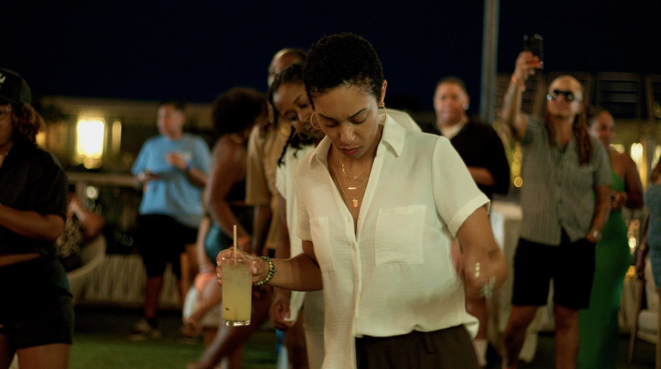 A woman with short curly hair wearing a white shirt and gold jewelry holds a pale yellow drink on a stick at a nighttime outdoor gathering with other people in the background.