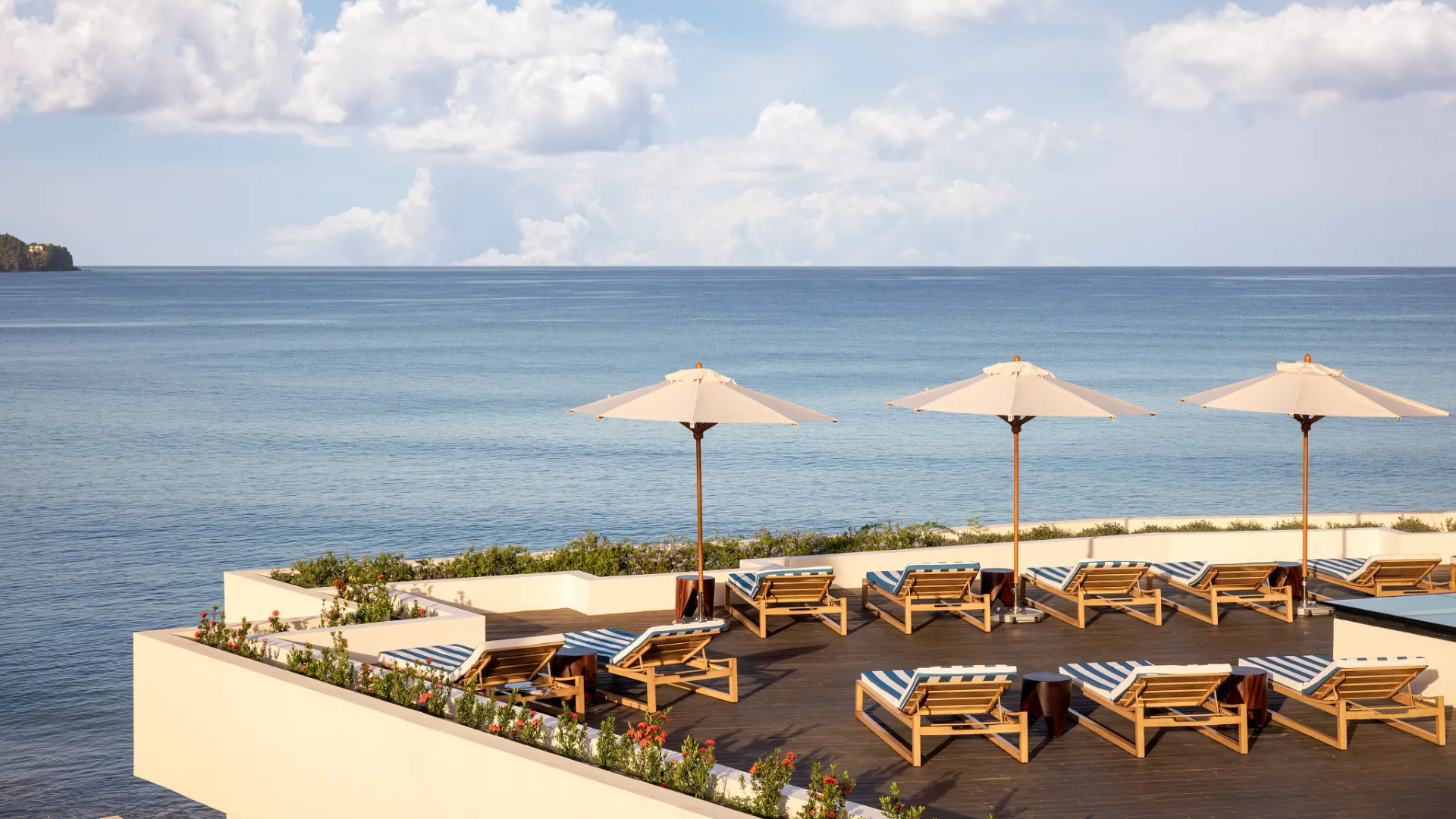 A rooftop lounge area overlooking the ocean with wooden lounge chairs with blue and white striped cushions, white umbrellas, and potted plants with flowers
