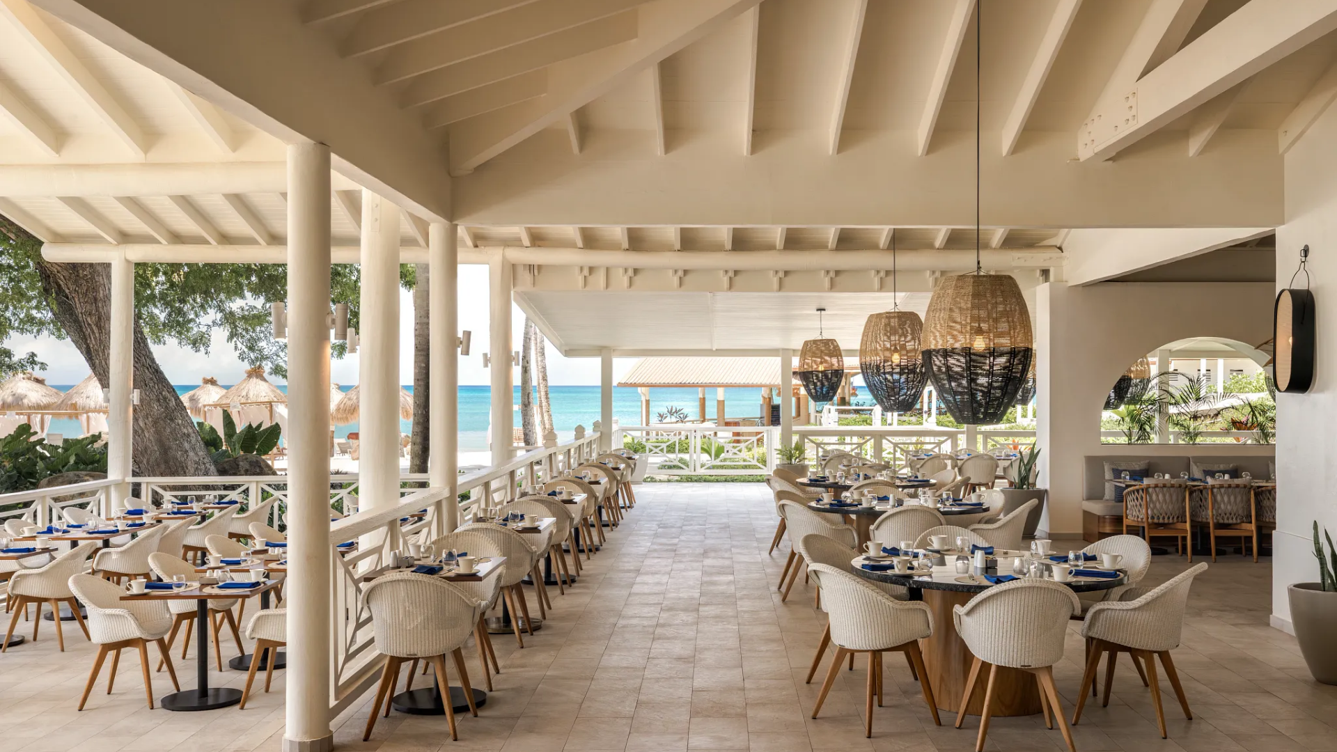 Beachside restaurant with white wicker chairs and tables set for dining, overlooking the ocean with straw umbrellas and trees in the background.