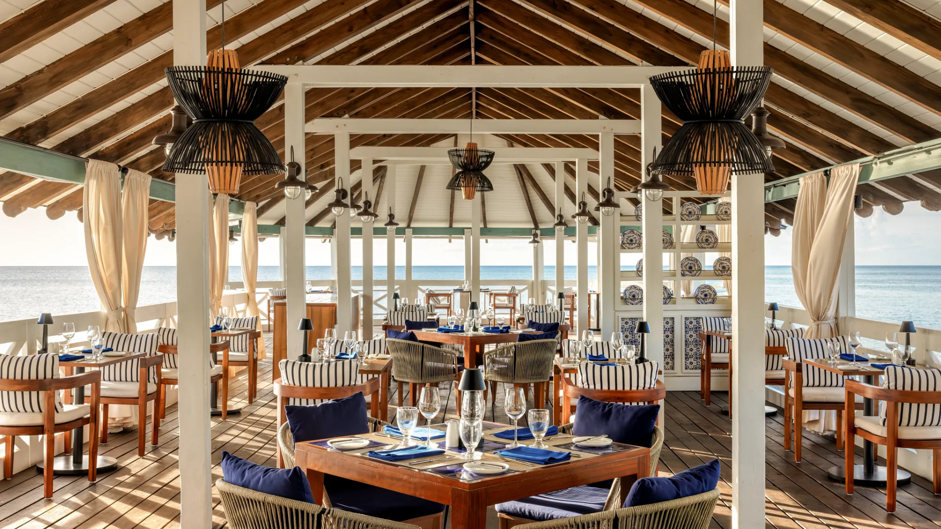 Indoor seaside restaurant with wooden floors, white pillars, and open sides overlooking the ocean. The ceiling has wooden beams and hanging black pendant lights. Dining tables are set with glasses, plates, and blue napkins, surrounded by wooden chairs with striped cushions.