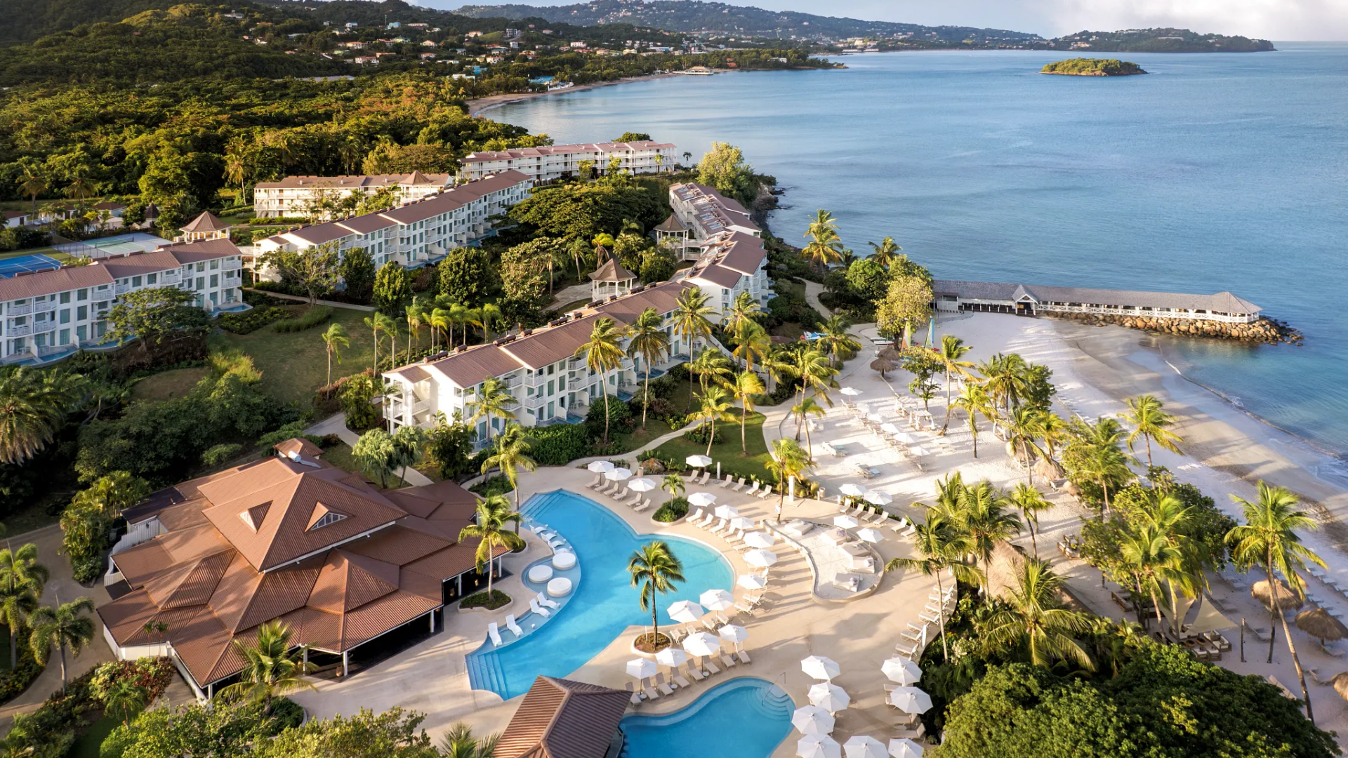 Aerial view of a tropical seaside resort with swimming pools, sandy beach, palm trees, and white apartment buildings along the shoreline.