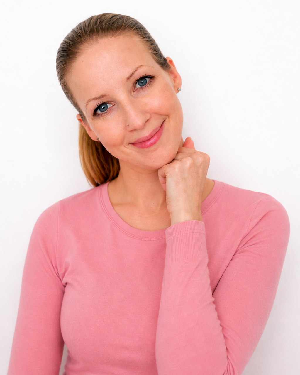 Nahaufnahme einer lächelnden Frau mit blauen Augen, braunen Haaren und rosa long-sleeve Top, die den Kopf neigt und die Faust unter dem Kiefer stützt.