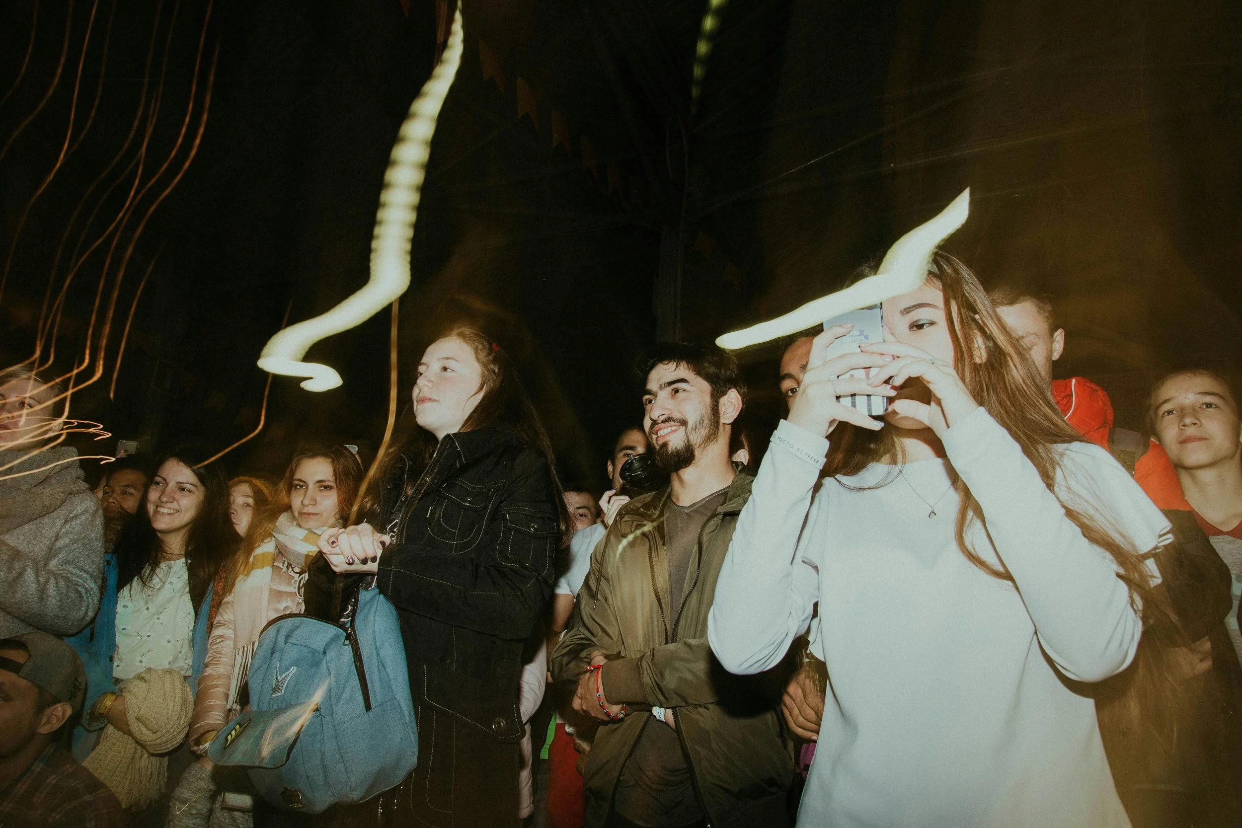 Gruppo di giovani durante un evento, alcune persone sorridono e qualcuna usa il cellulare, in un ambiente con luci soffuse e movimento.