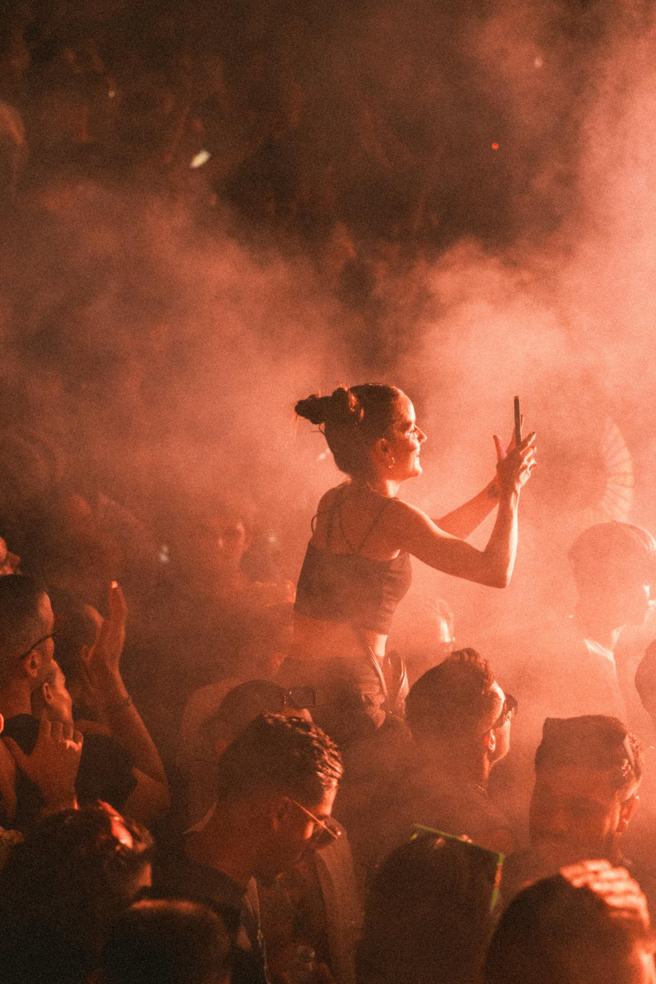 Una donna si trova in mezzo a un pubblico in un ambiente affollato e fumoso, probabilmente a un concerto o a un evento musicale, mentre prende un selfie con il cellulare.
