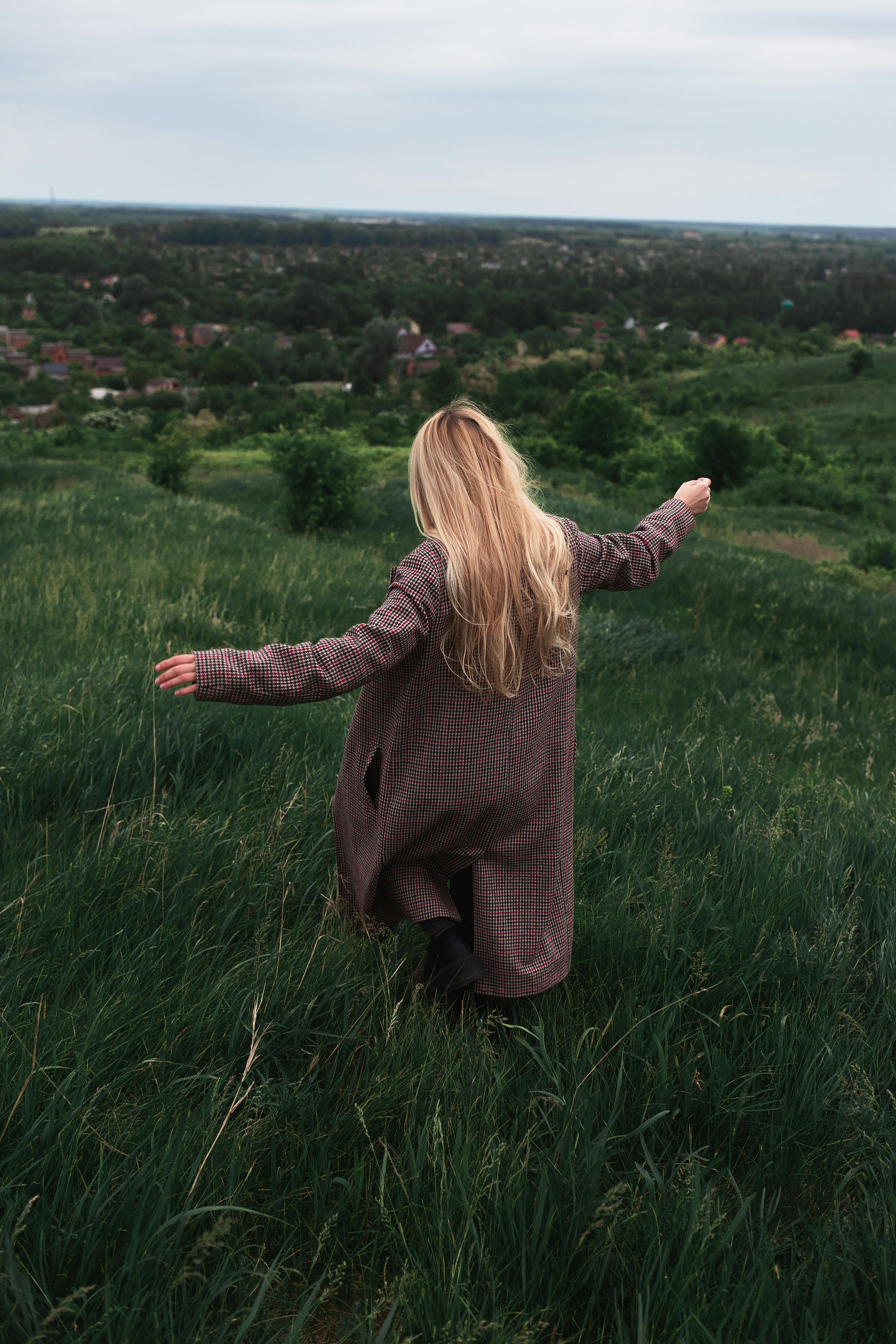 Una donna con capelli biondi lunghi, indossa un cappotto a quadri e si trova in un prato verde, con le braccia aperte, che guarda un paesaggio di campagna con case e alberi sotto un cielo nuvoloso.