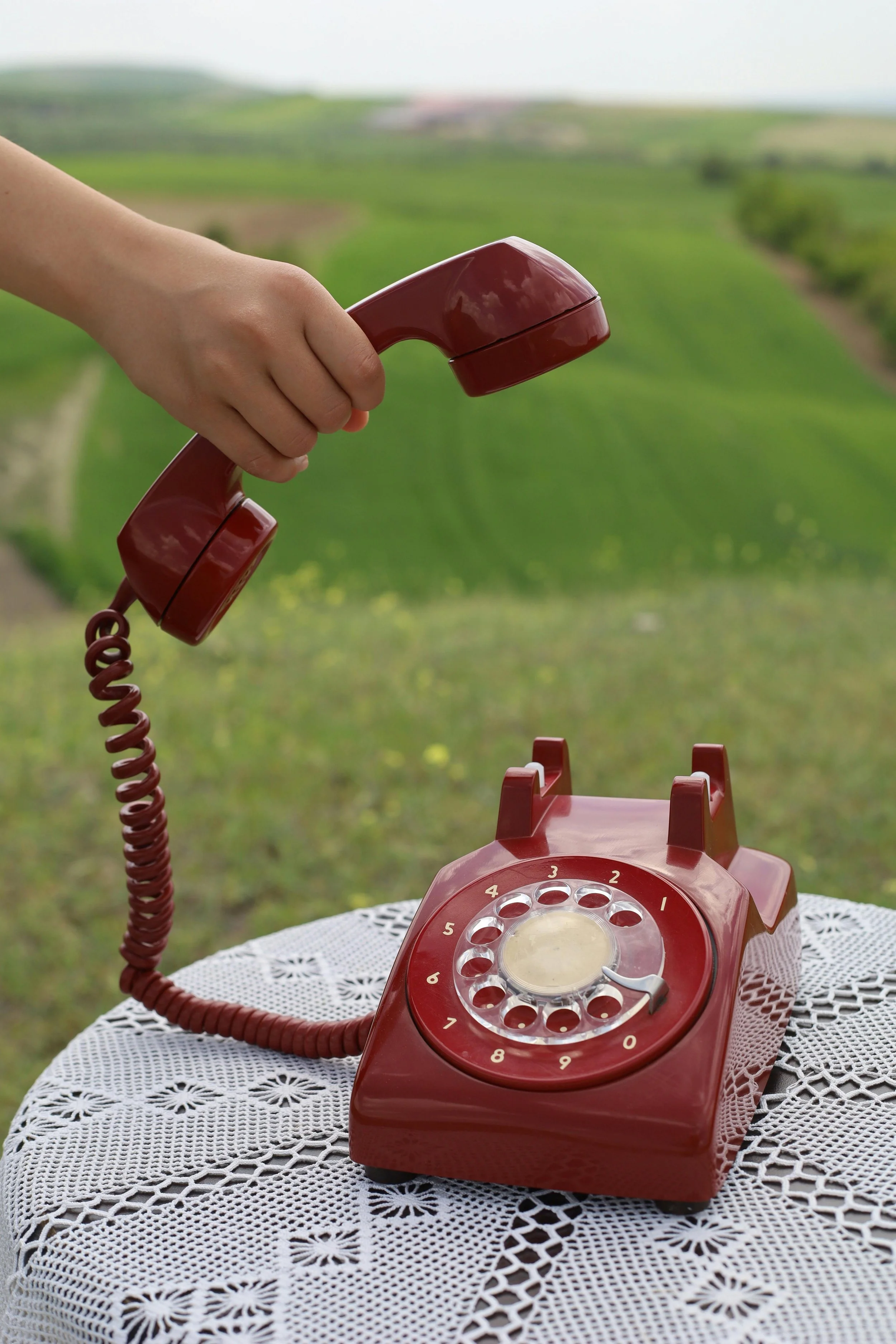 Telefono vintage rosso su tavolo di pizzo bianco, con sfondo di campagna verde e colline.