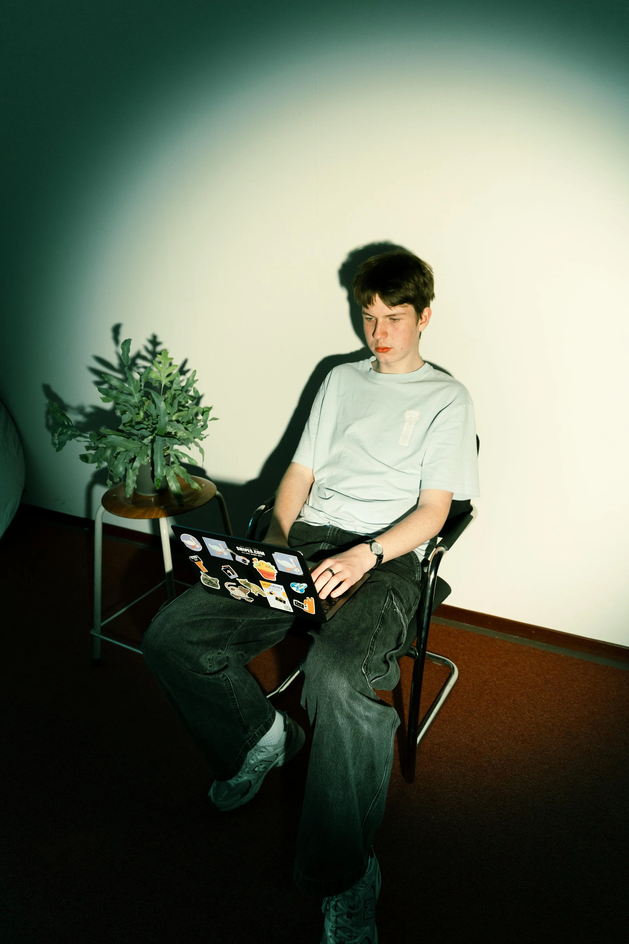 Ragazzo seduto su una sedia con un laptop sul suo grembo, indossa una maglietta grigia e pantaloni scuri, accanto a una pianta in un vaso.