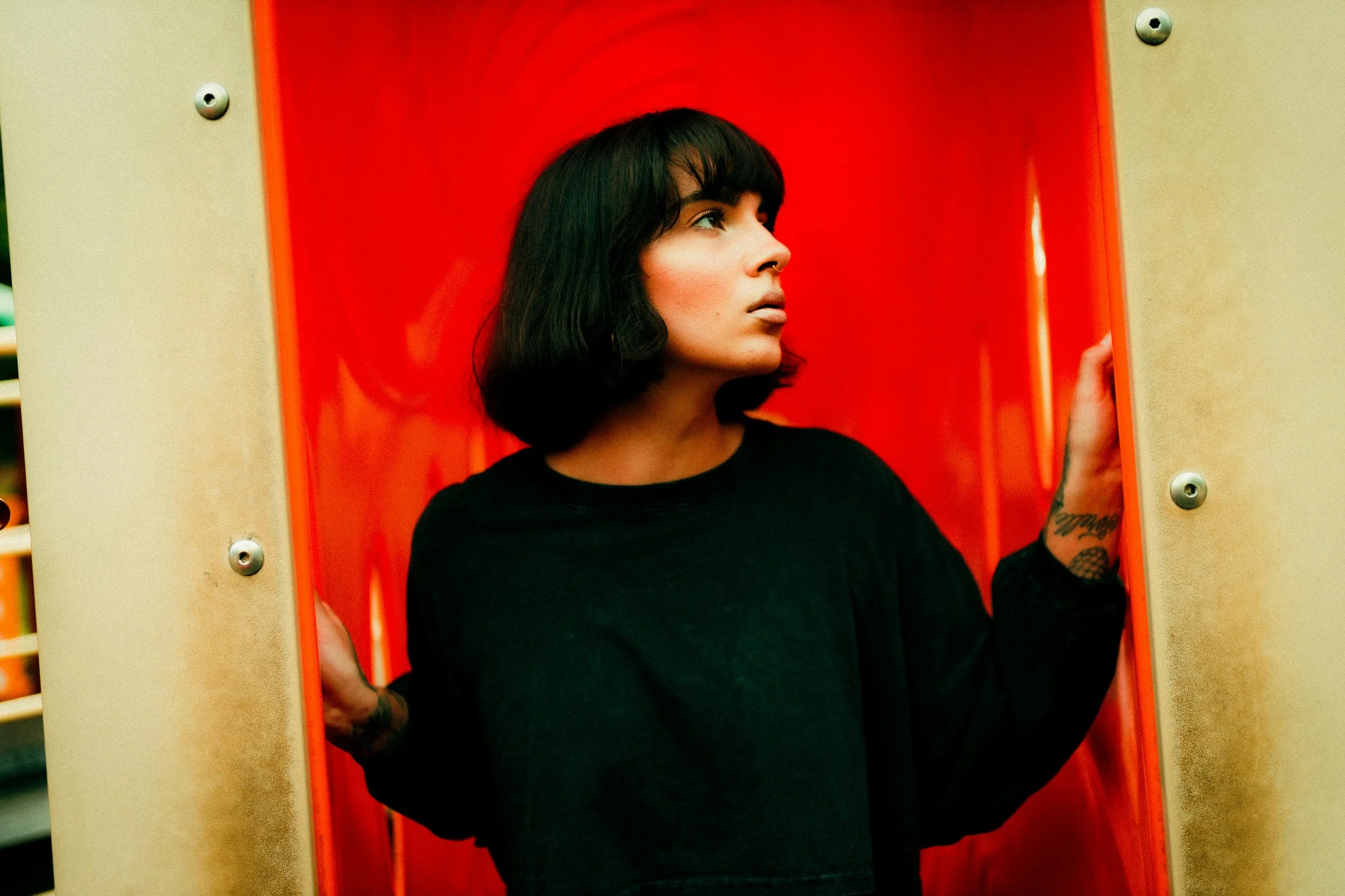 Una giovane donna con capelli neri a spazzola, maglietta nera, in piedi in una cabina rossa. Con tatuaggi visibili sulle mani, guarda verso l'alto a sinistra.