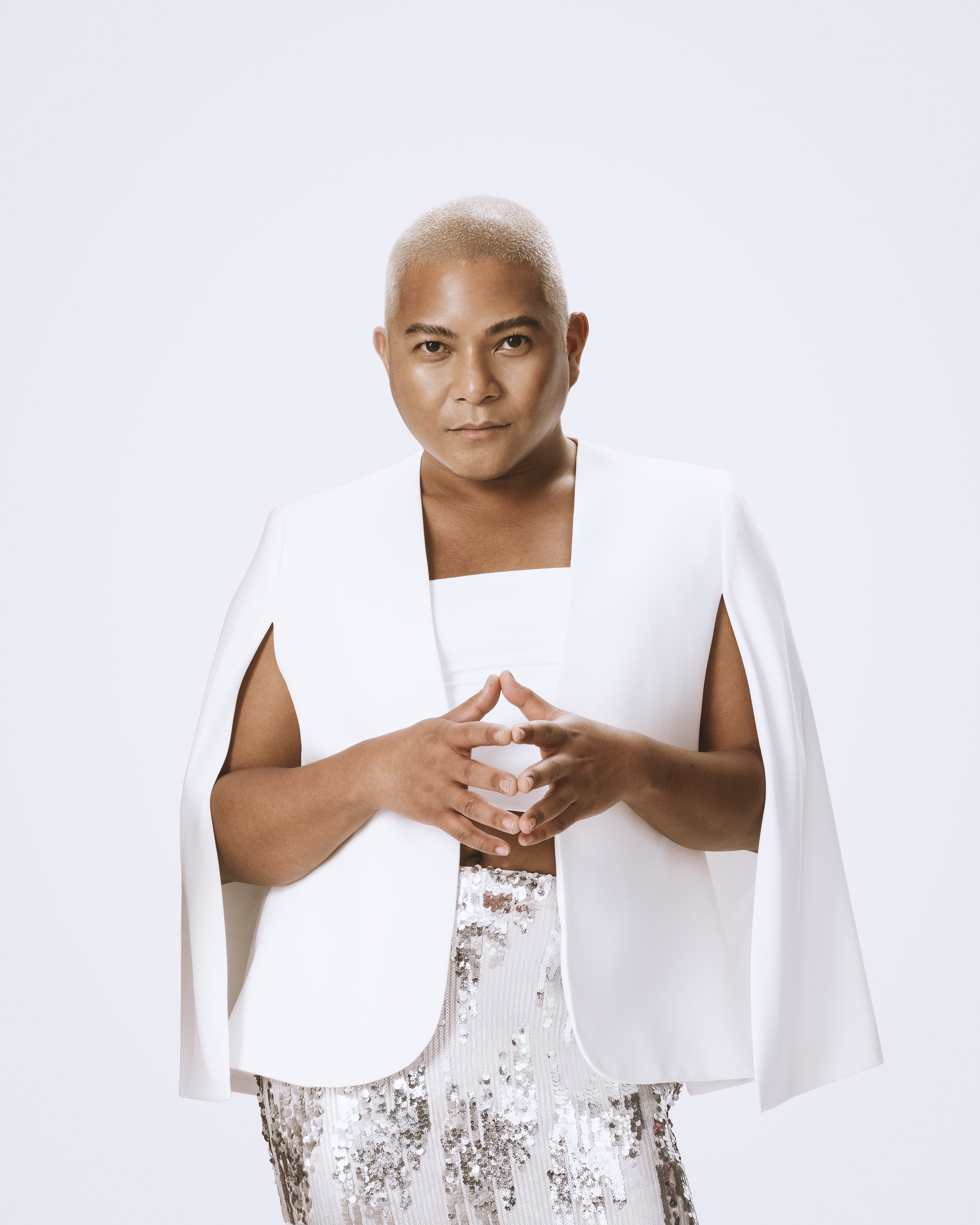 Domingo @mydomingo Lumanog profile image gay man in a white cape, blonde hair, long lashes from sugar lash pro, hands and fingers aligned looking empowered and strong