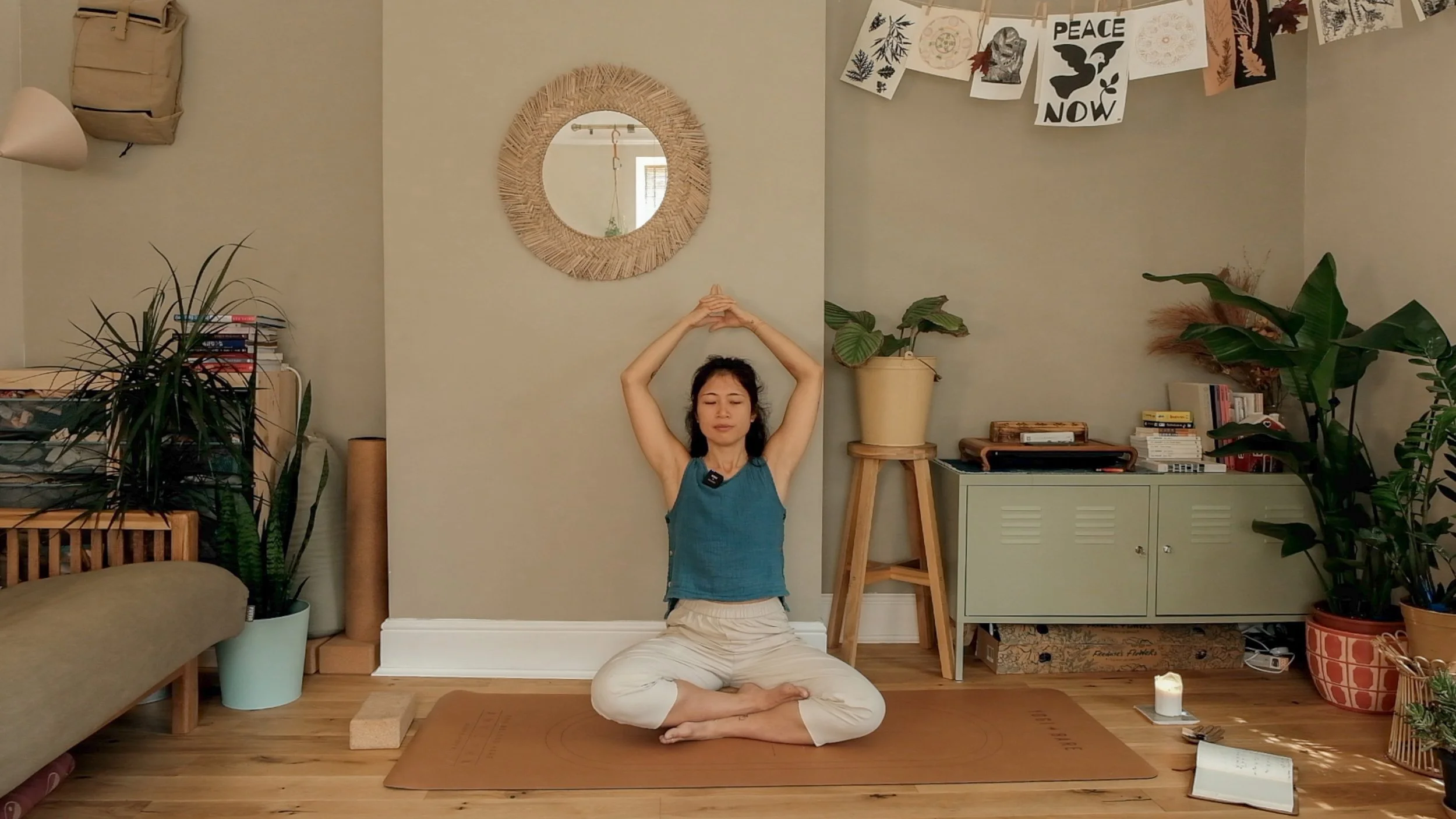 A woman practicing yoga in a cozy, plant-filled room. She sits cross-legged on a yoga mat with her arms raised above her head. The room features a mirror, potted plants, a bookshelf, and hanging artwork.