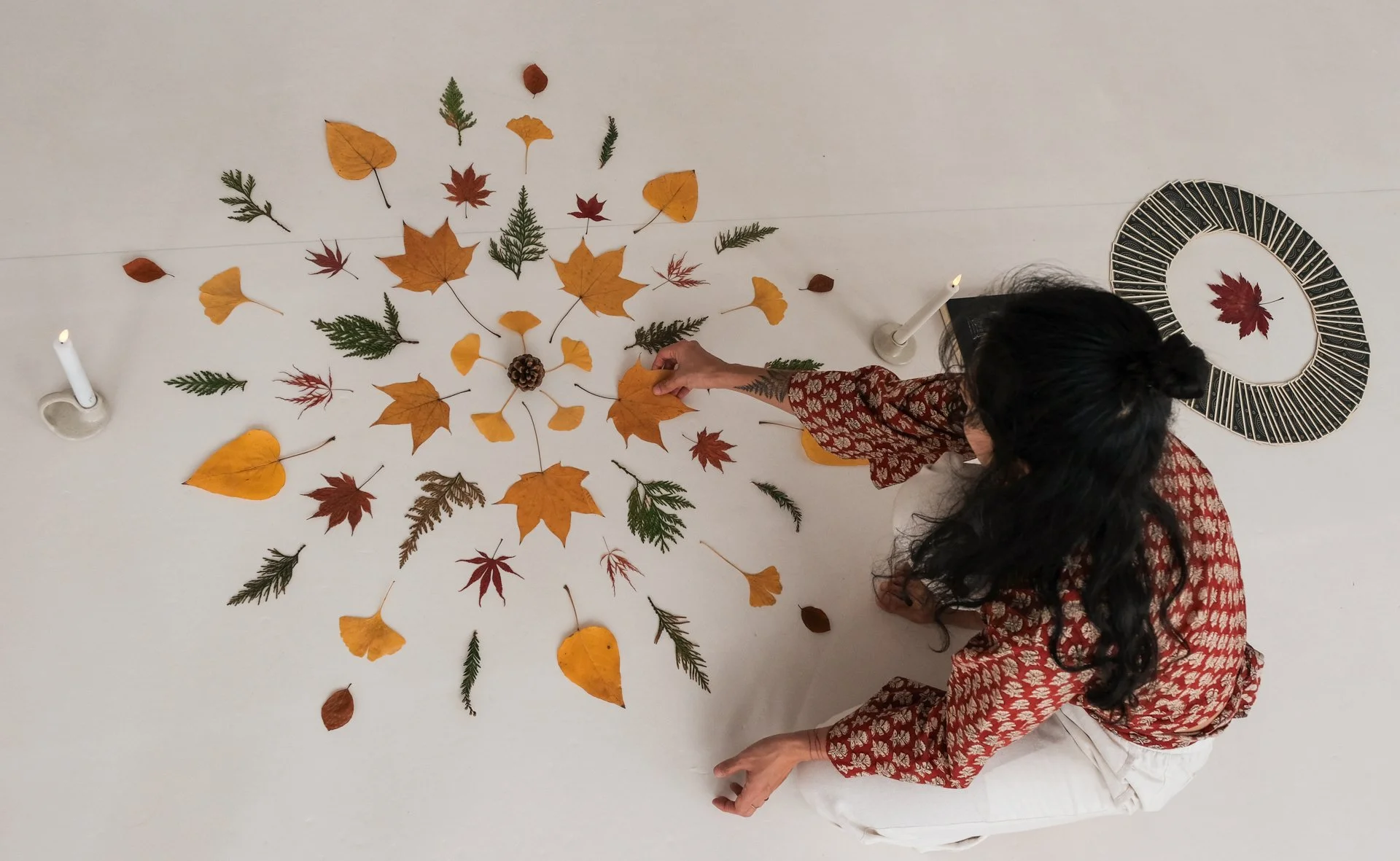 A woman in a patterned red top arranging fall leaves and a pine cone on a white floor, with candles and a decorative circular object nearby.