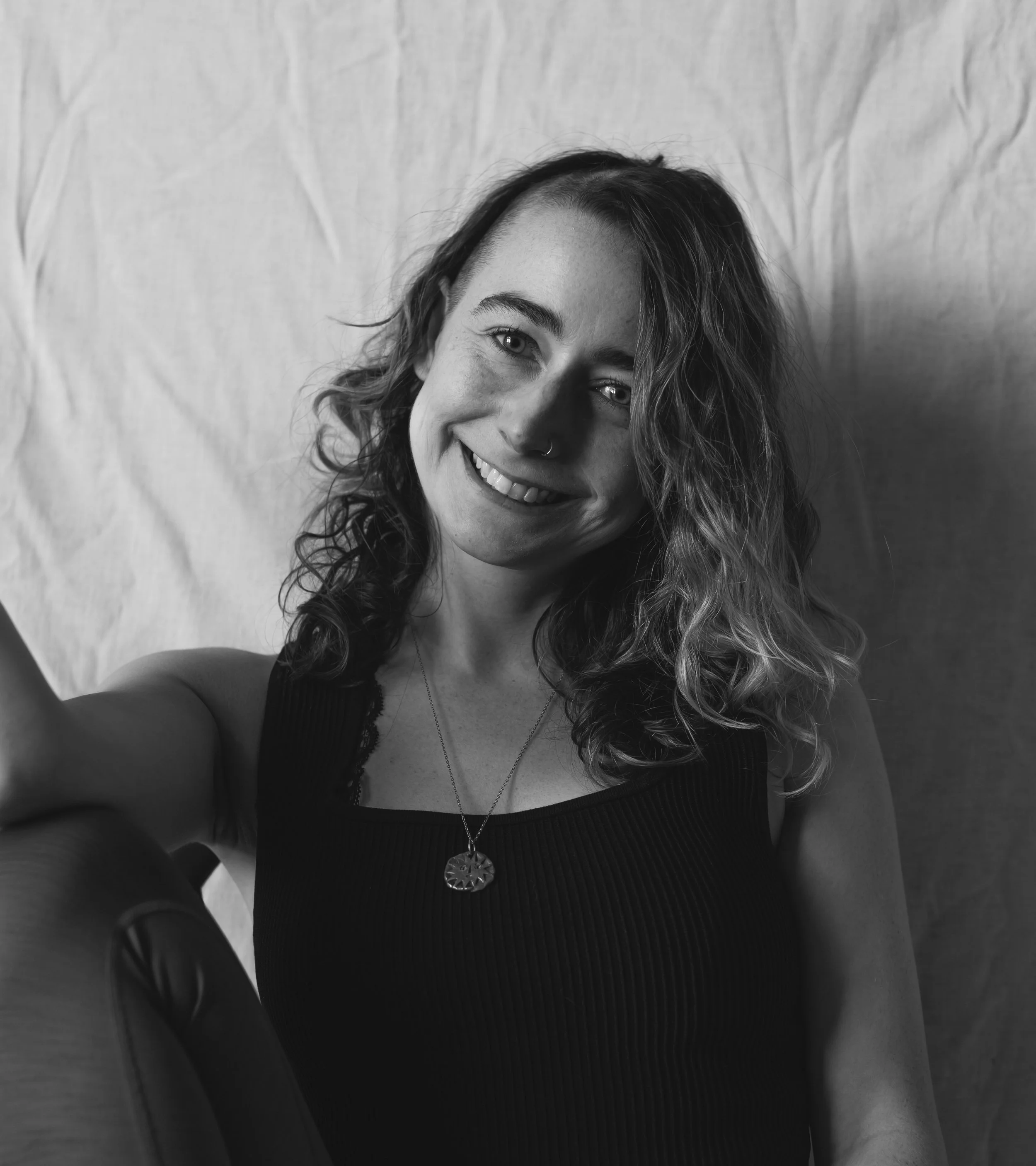 A smiling woman with wavy hair taking a selfie in black and white, sitting in front of a fabric backdrop.