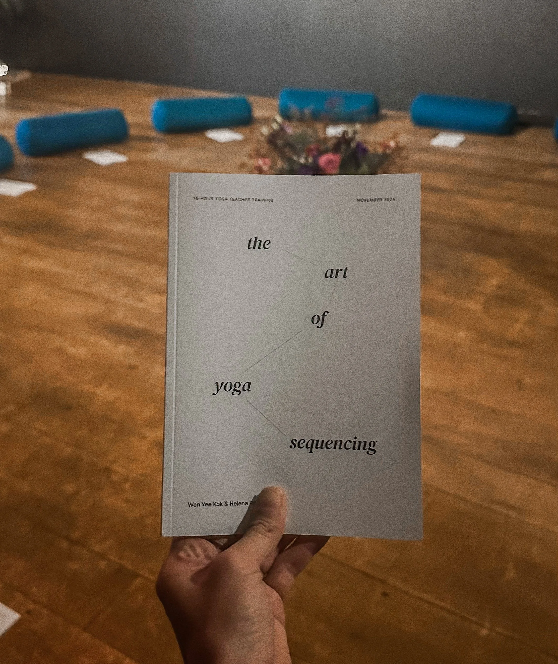 A person holding a book titled 'the art of yoga sequencing' in a room with a wooden floor and chairs with blue covers in the background.