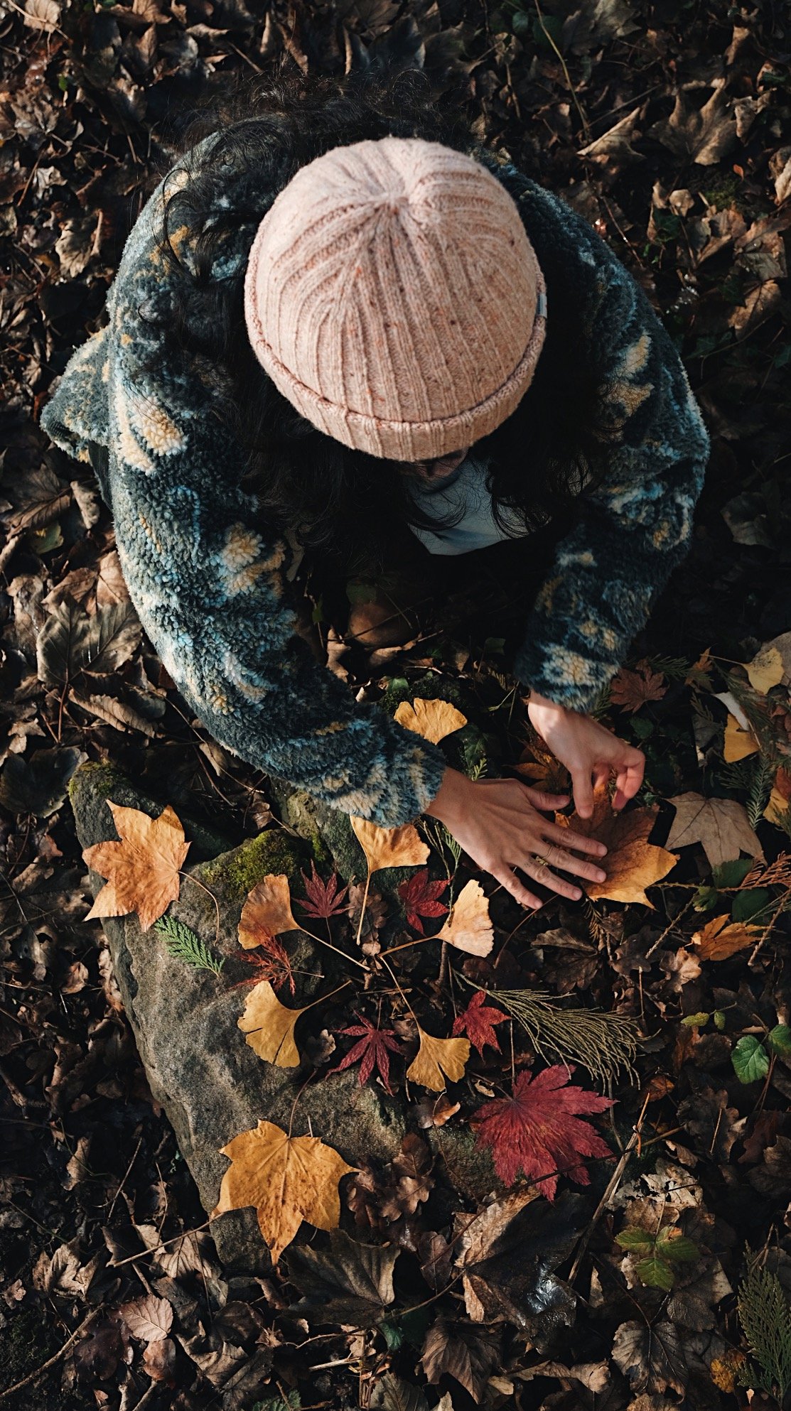 A person with curly hair wearing a peach knit beanie and a patterned fleece jacket, crouching among fallen autumn leaves on the forest floor.