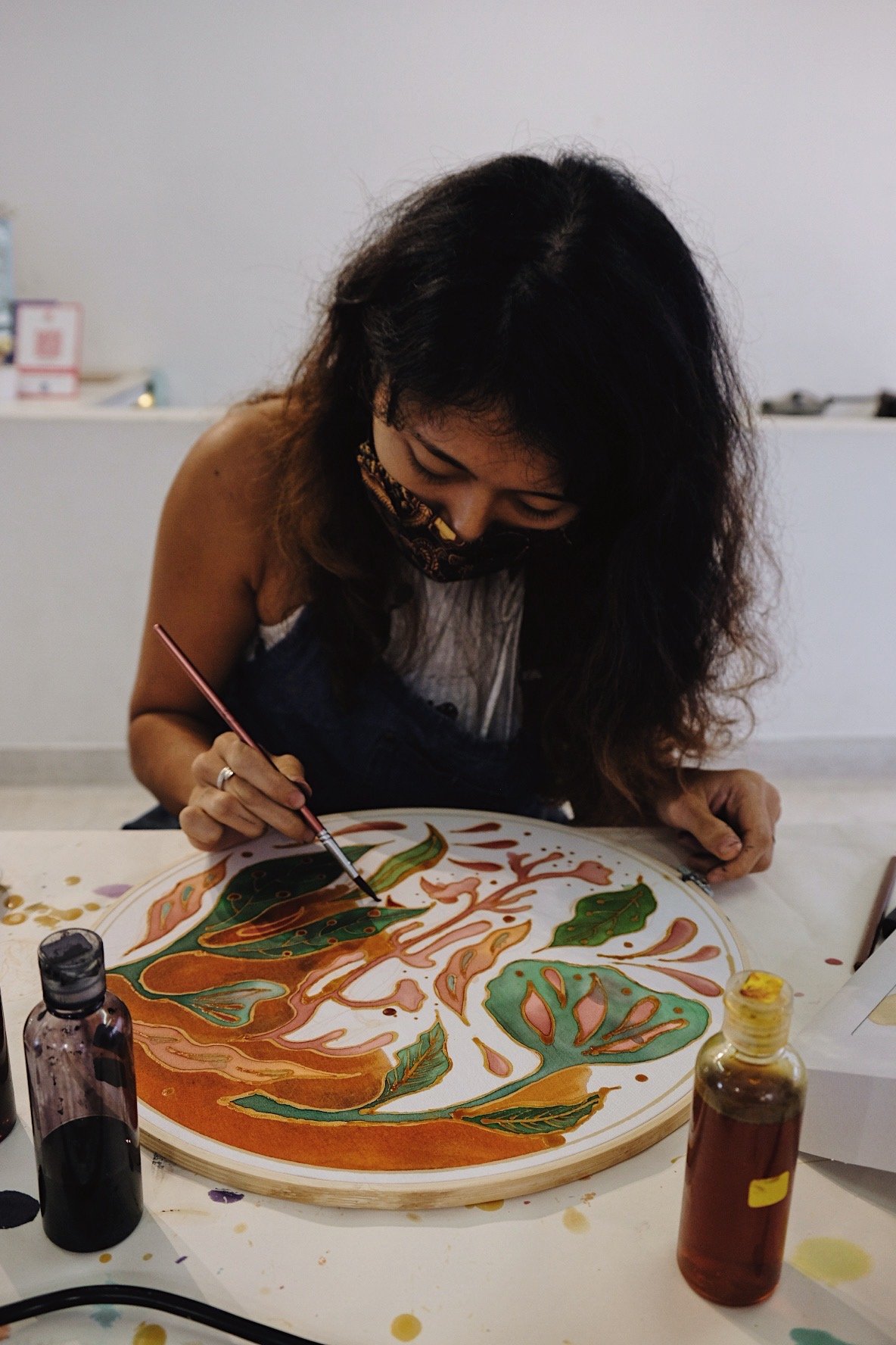 Person painting detailed artwork with brush on circular canvas, surrounded by art supplies.