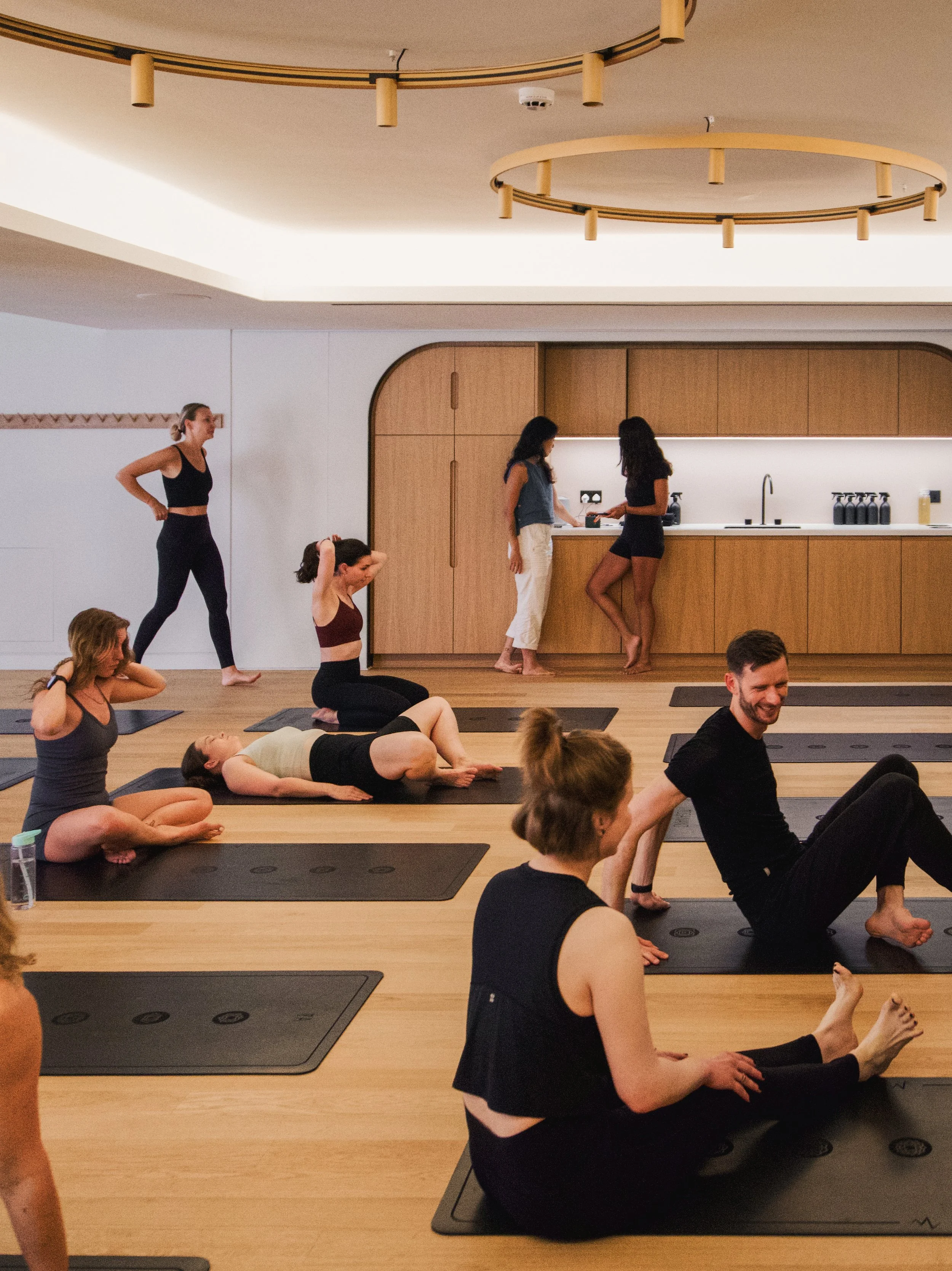 People participating in a yoga class in a modern studio with wooden flooring, black mats, and a wooden kitchenette in the background.