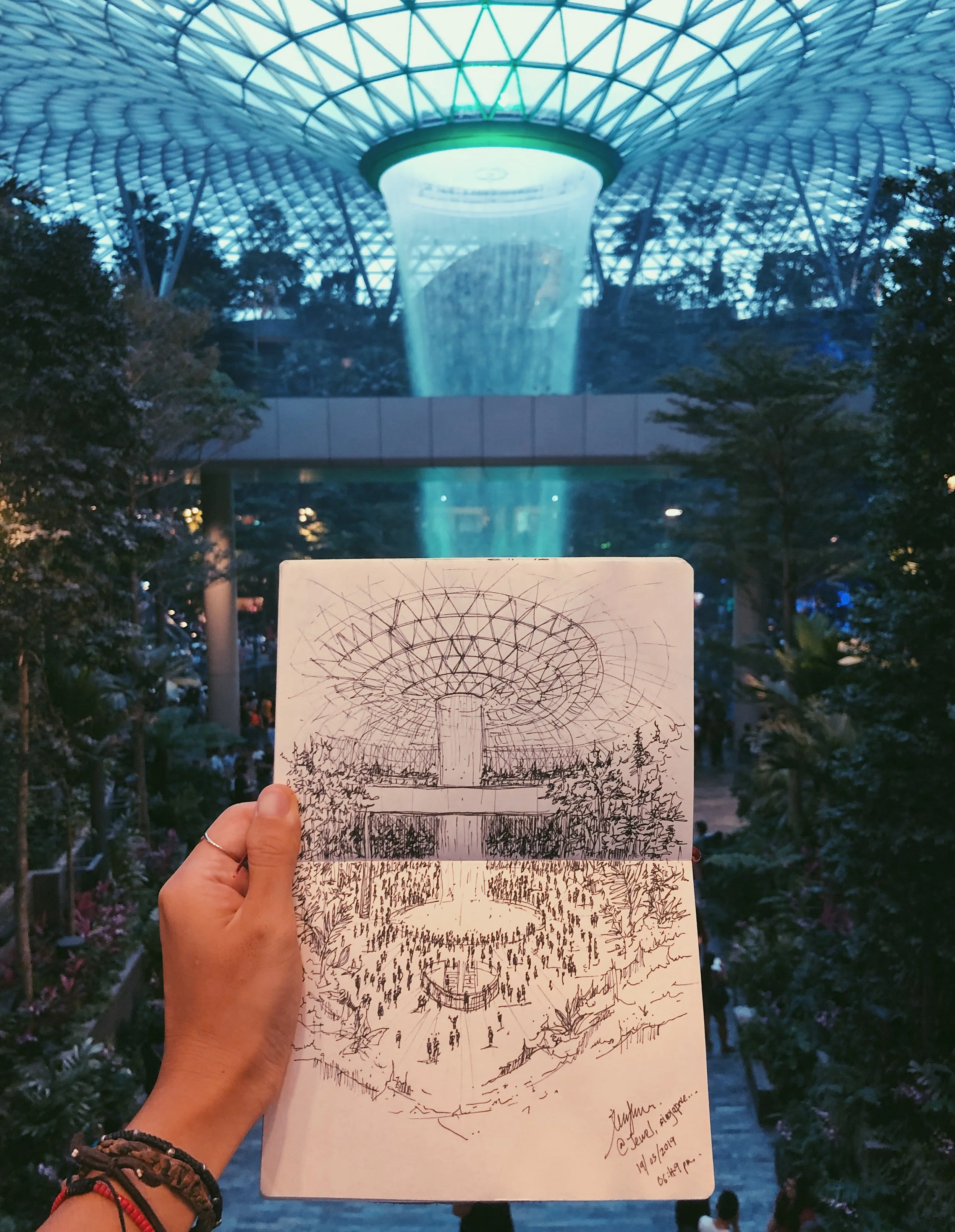 A person holds a sketchbook featuring a drawing of a large indoor waterfall and dome ceiling, matching the real scene in the background at Jewel Changi Airport, Singapore.