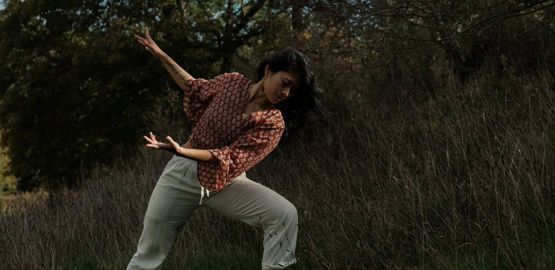 A woman with long dark hair wearing a red patterned blouse and light-colored pants is dancing outdoors in a grassy field with trees in the background.