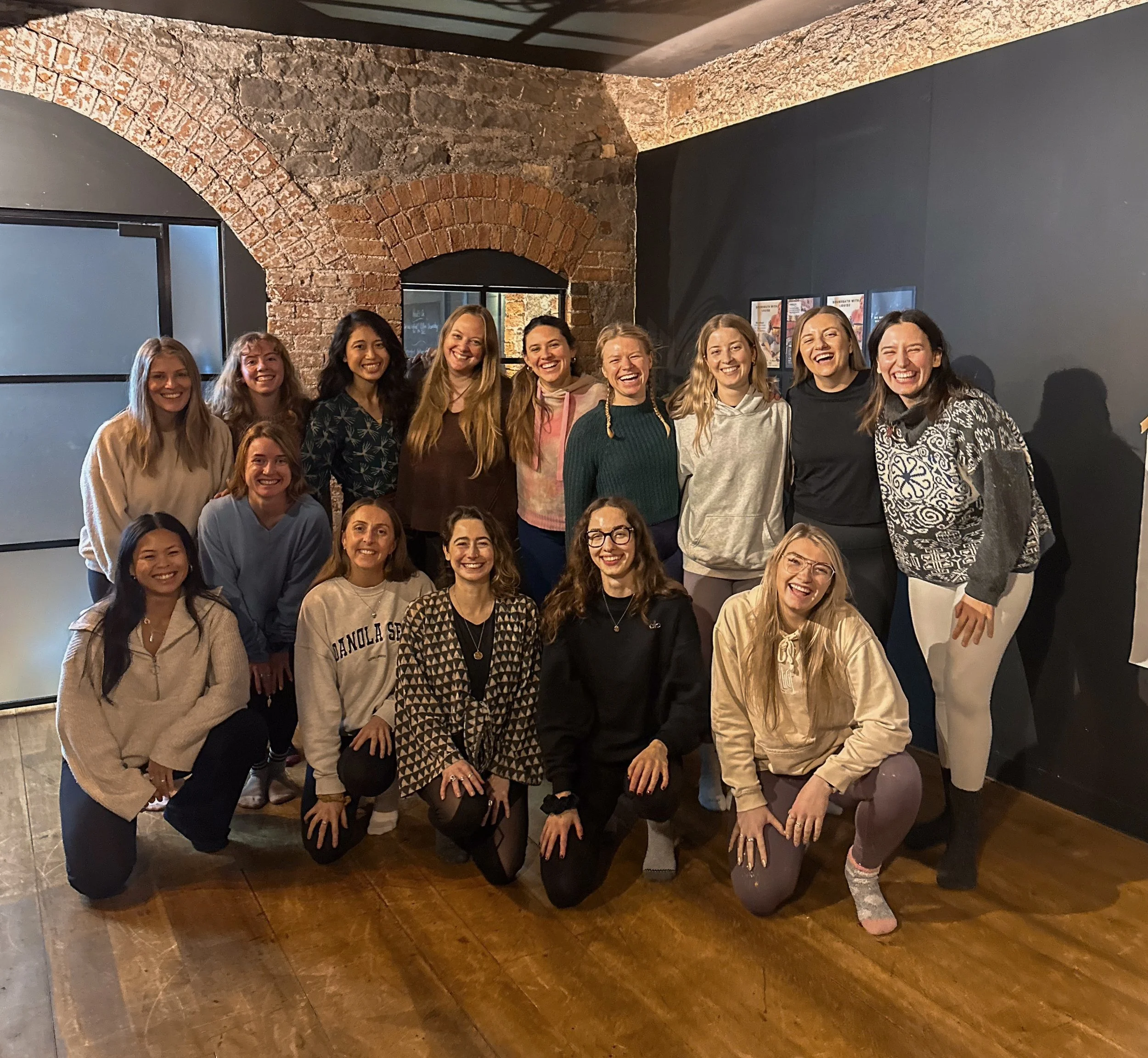 A group of 15 women smiling and posing together indoors in front of a brick wall and black wall with framed pictures, some women are kneeling in front and others standing behind.