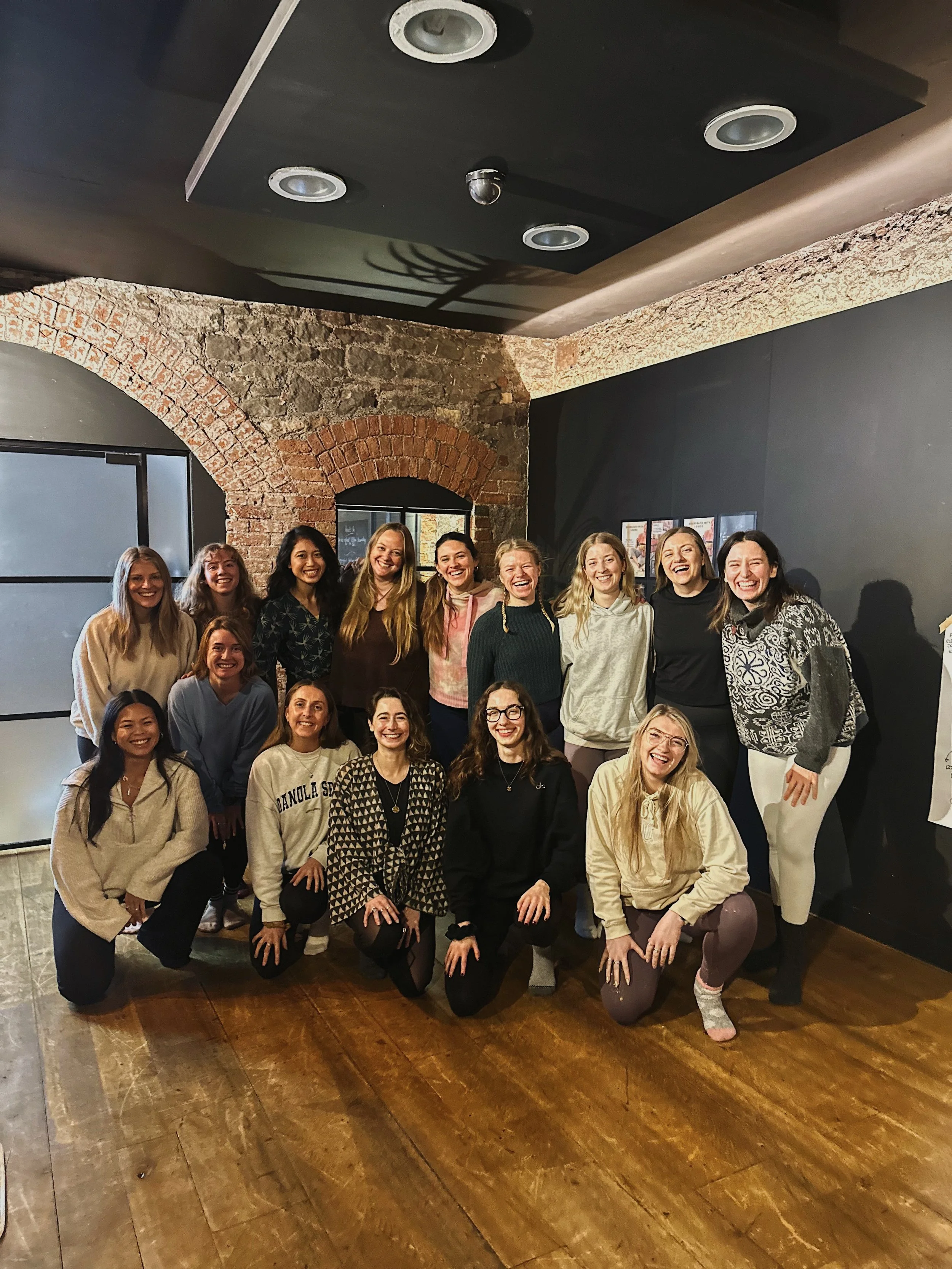 Group of women posing indoors on a wooden floor with brick and dark walls