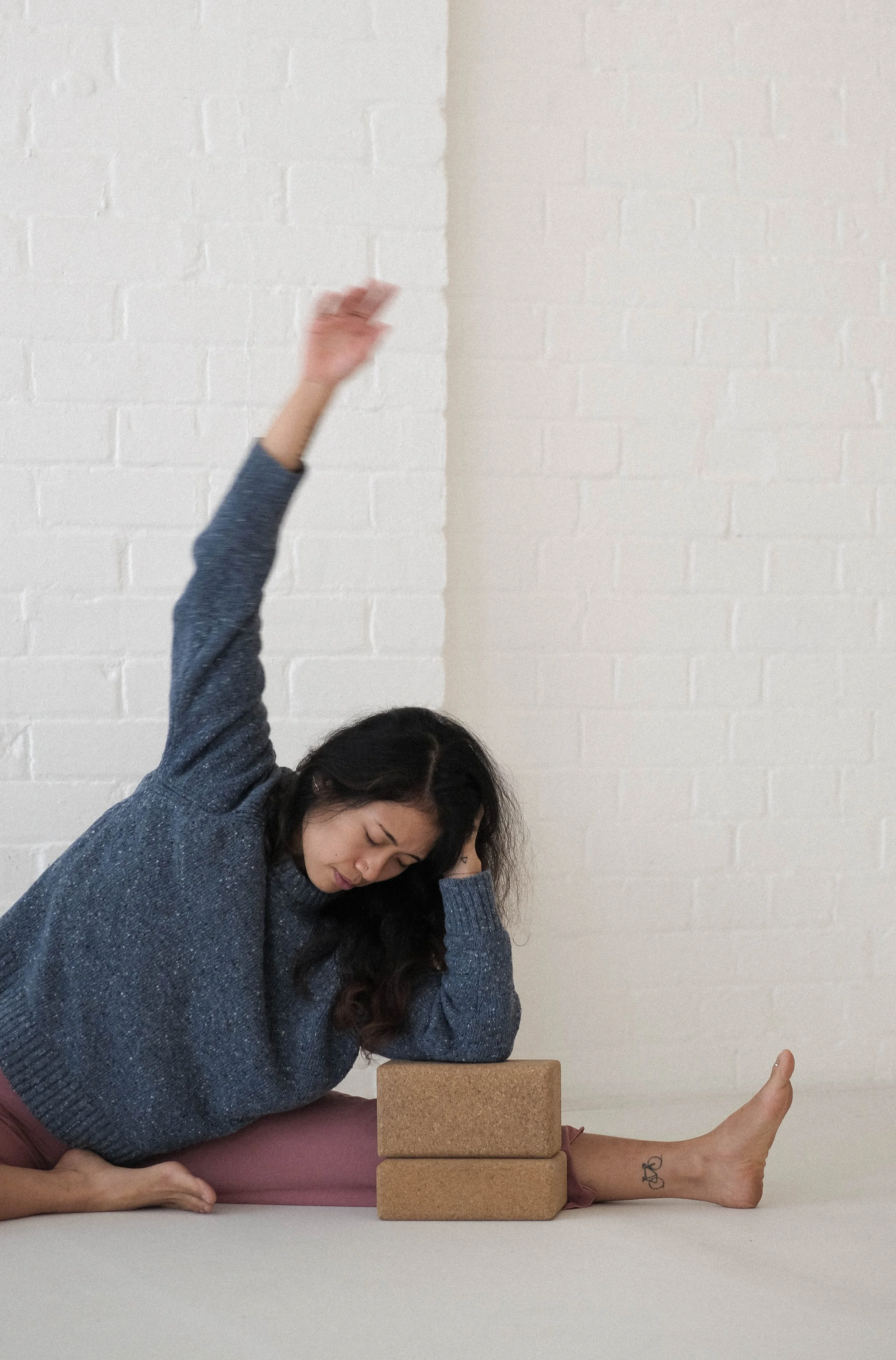 Person in a seated yoga pose stretching with arm raised, next to two yoga blocks, against a white brick wall.