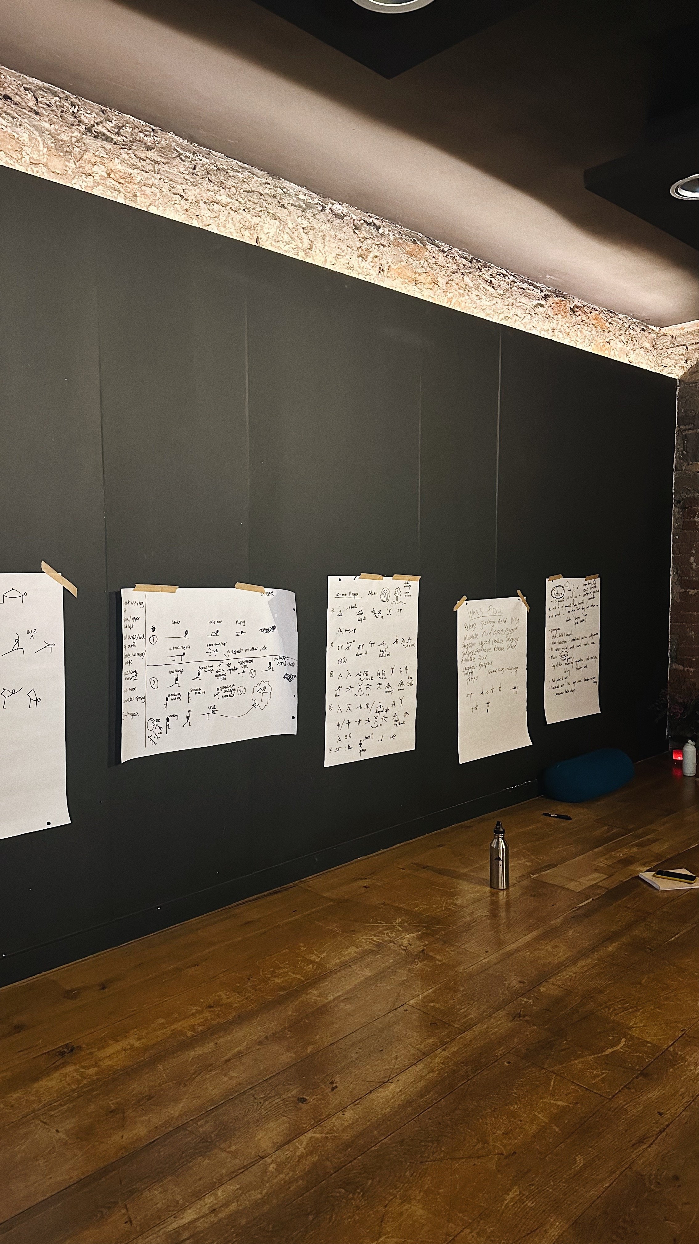 Room with dark walls and wooden floor, featuring large sheets of paper with drawings and notes taped to the wall. A stainless steel water bottle and a yoga bolster are on the floor.