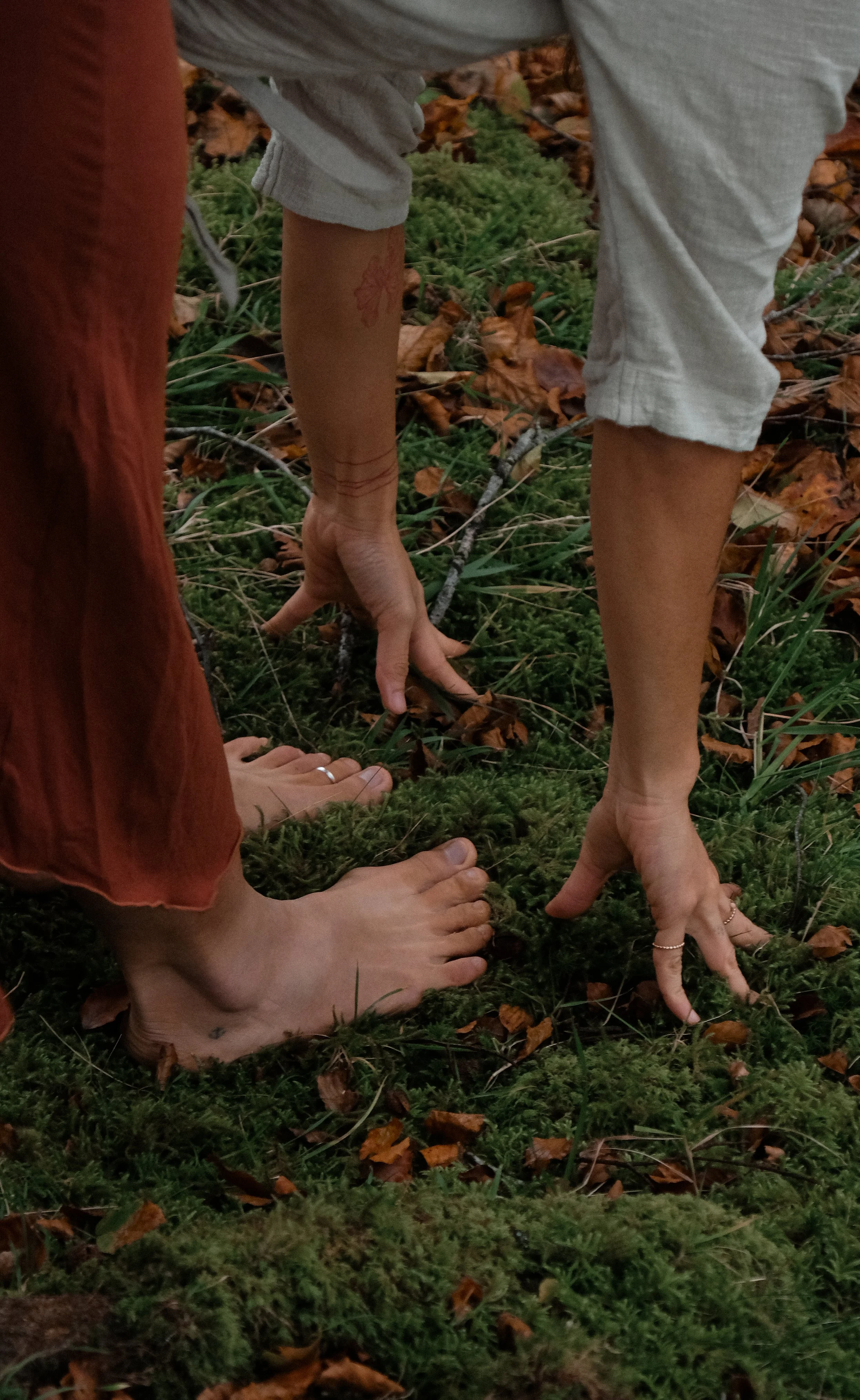 Barefoot people standing on mossy ground with fallen leaves; hands touching the ground.