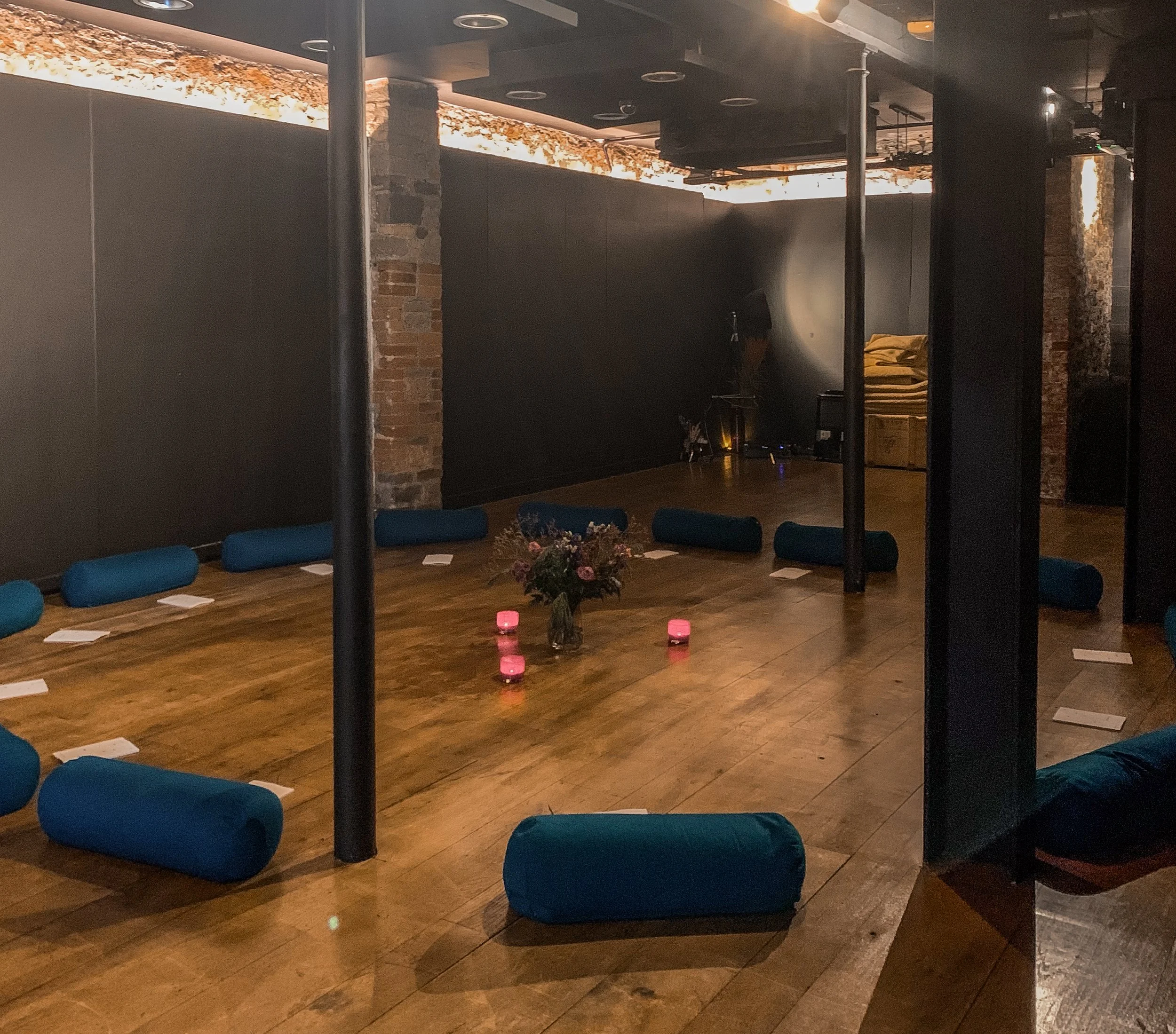 A dimly lit meditation or yoga space with a central bouquet of flowers surrounded by four pink candles. Blue cylindrical bolster pillows are arranged around the perimeter on a wooden floor, with some white papers placed next to each one. The room has