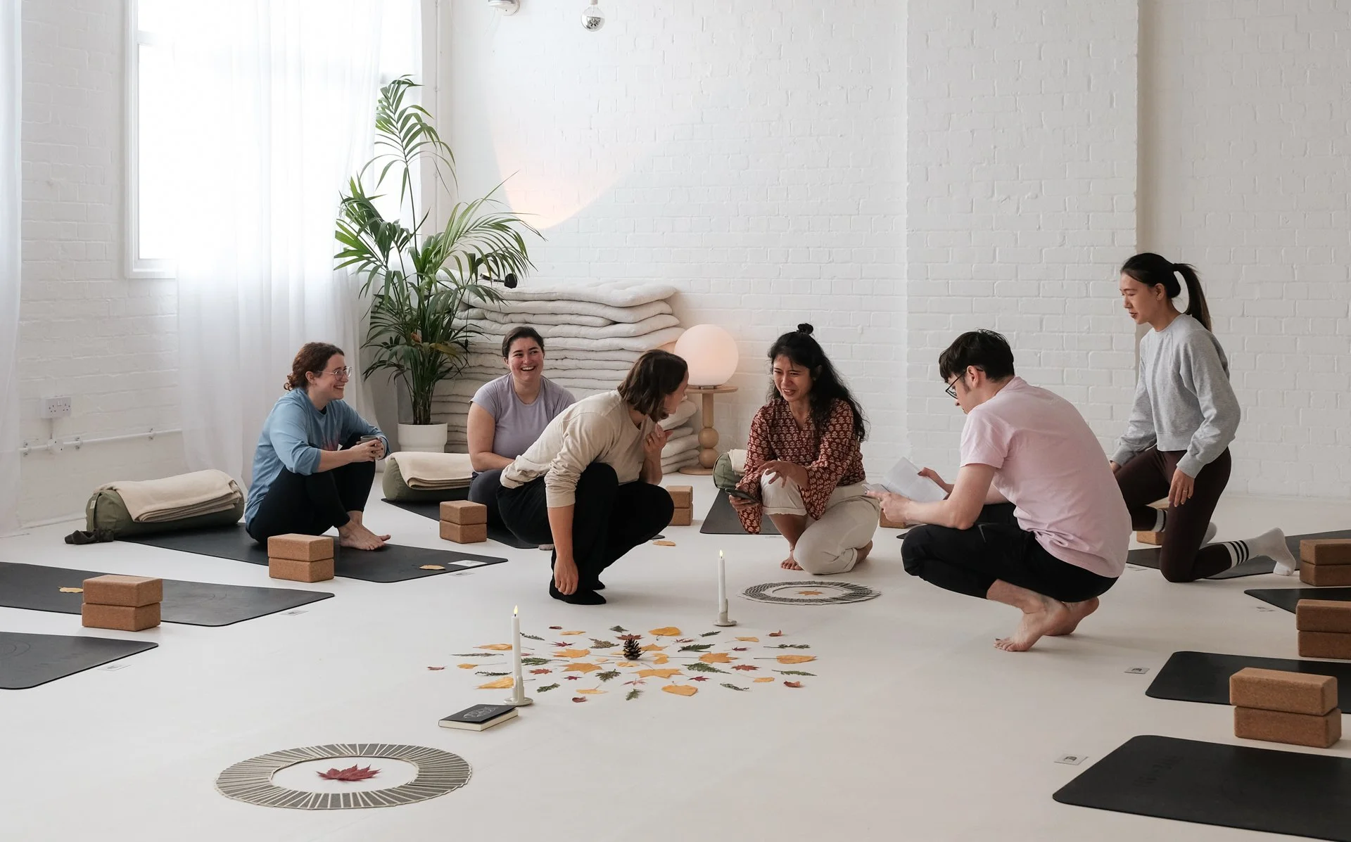 Group of people participating in a yoga or meditation session in a bright, minimalist room with white walls, plants, and natural light.