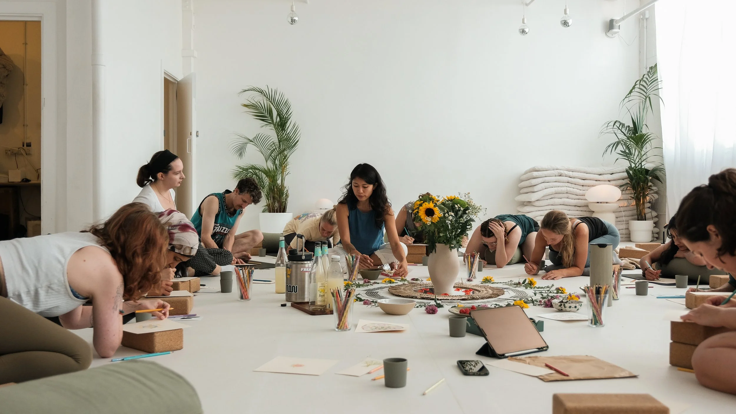 Group of women participating in a yoga or meditation class in a bright, minimalist room with potted plants and tied blankets in the background. They are sitting on the floor around a central decorative arrangement with flowers, drawing or writing.