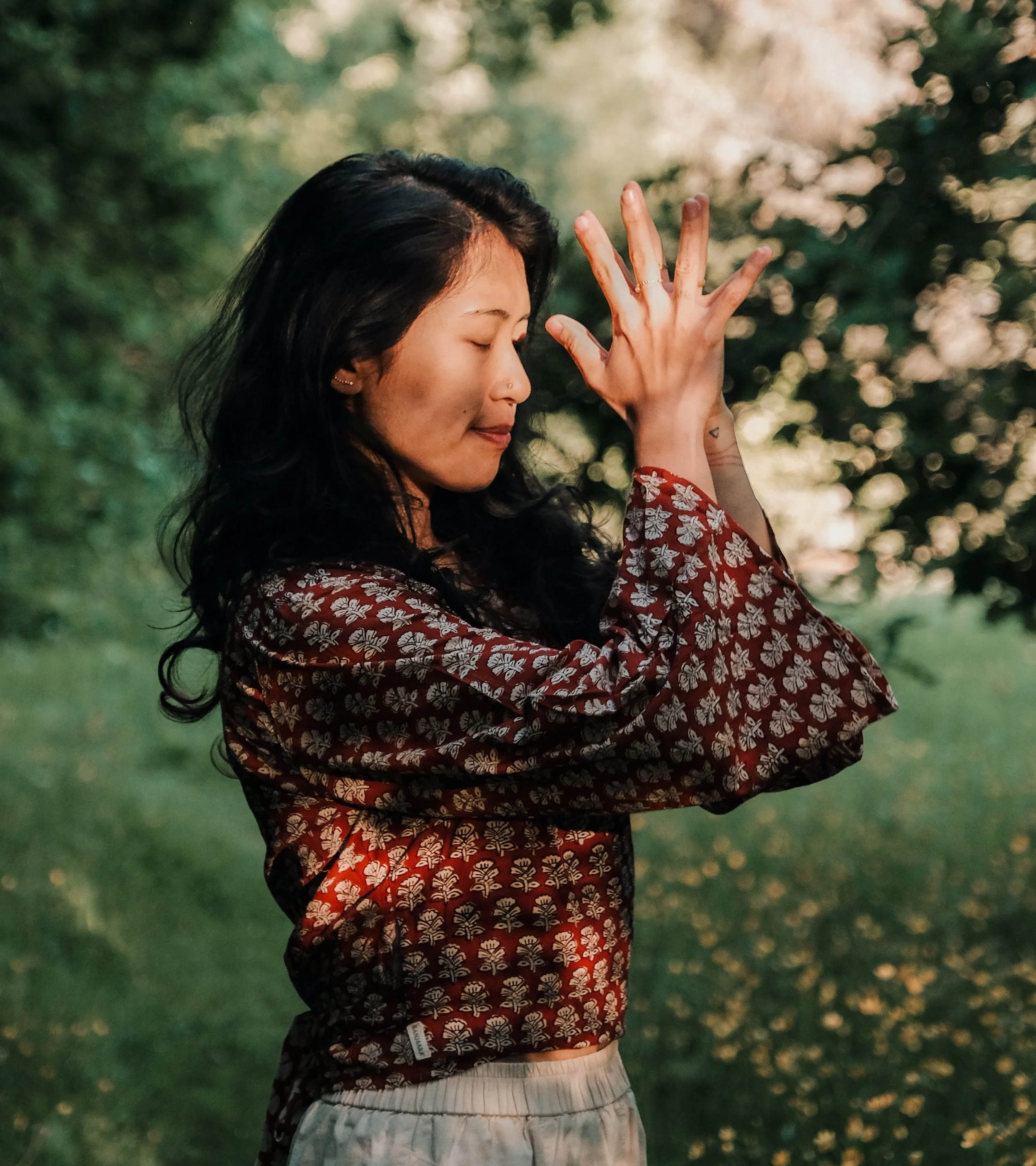 A woman with long dark hair in a patterned blouse and light-colored pants with closed eyes, holding her hand near her face in a peaceful outdoor setting.