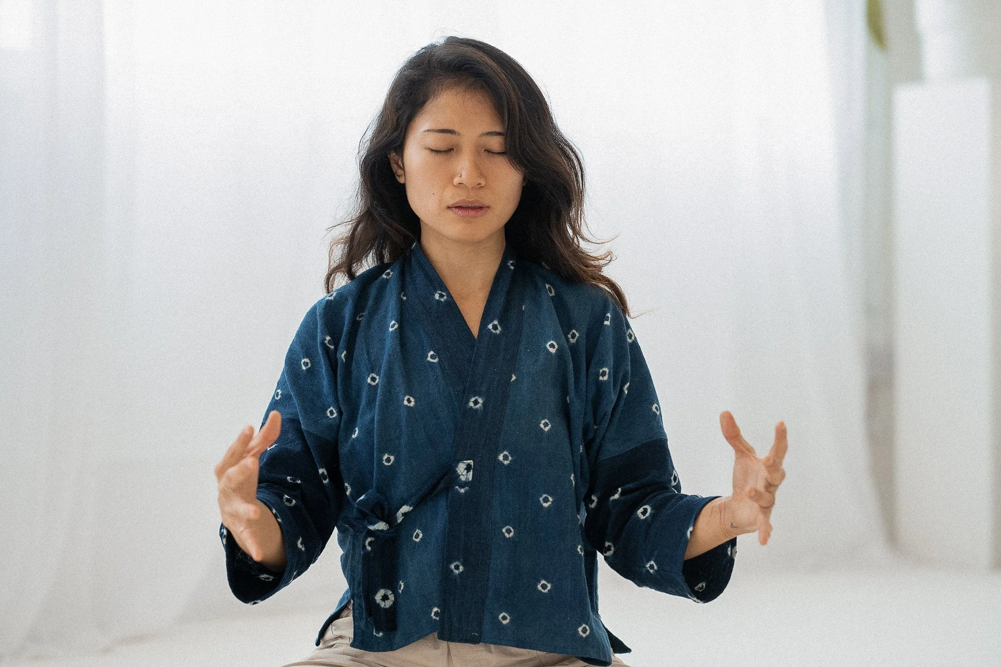 Woman meditating with closed eyes, wearing a blue patterned robe, sitting in a calm, light-filled space.