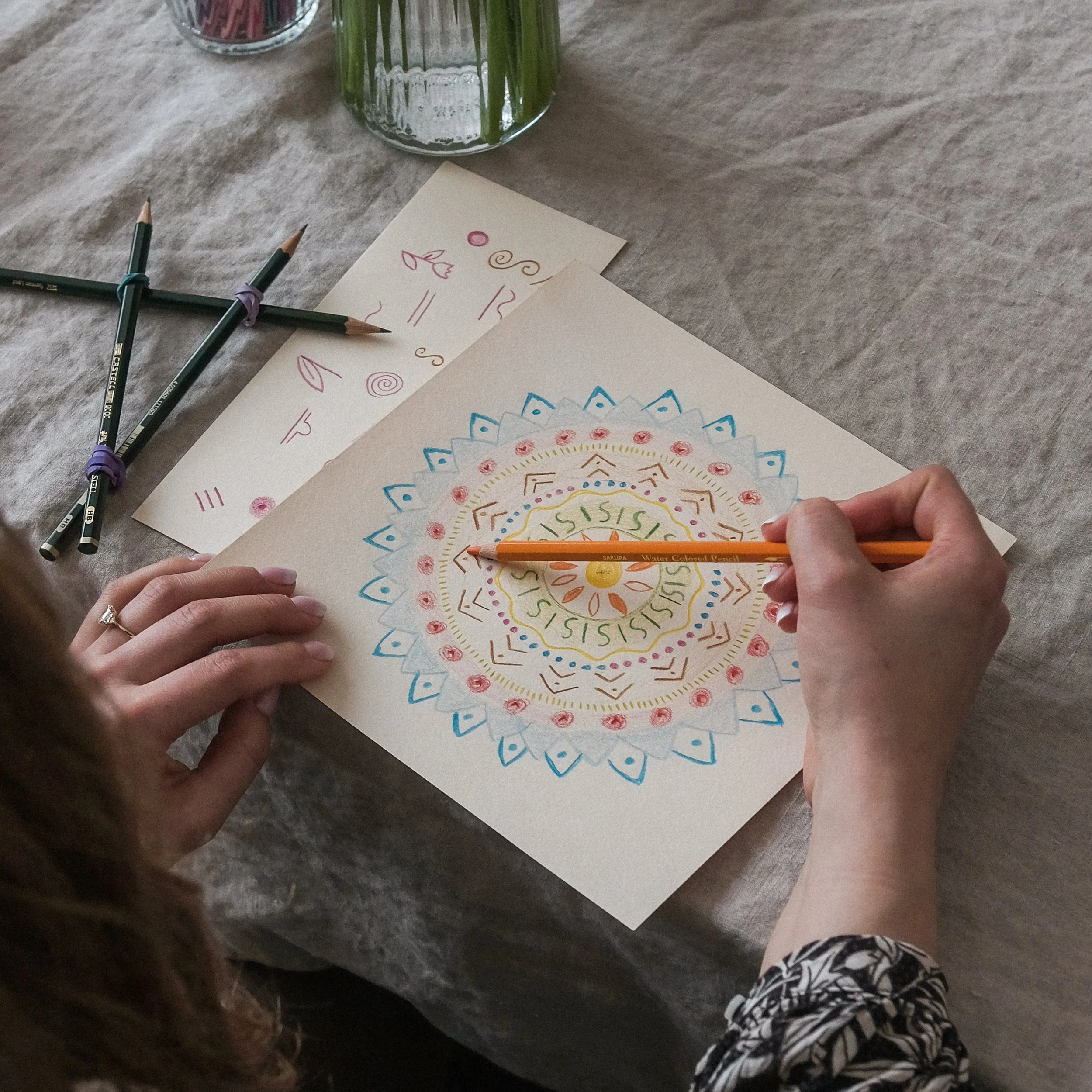 Person coloring a mandala design with colored pencils on a piece of paper, with additional colored sketches nearby, on a table with a tablecloth.