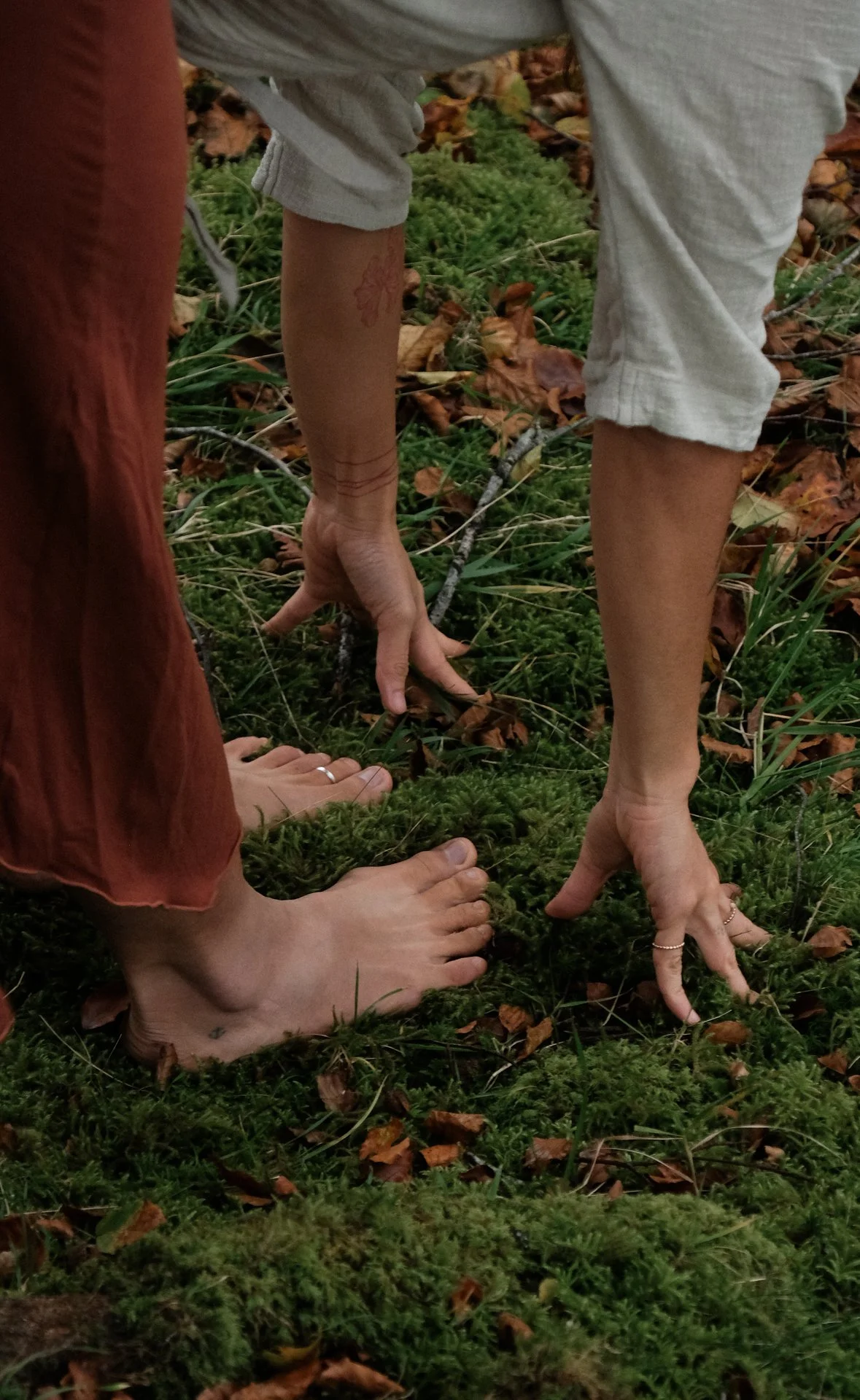 Two people are barefoot in a mossy, leafy forest, with one person kneeling and touching the ground while the other stands and points to a stick in the moss.