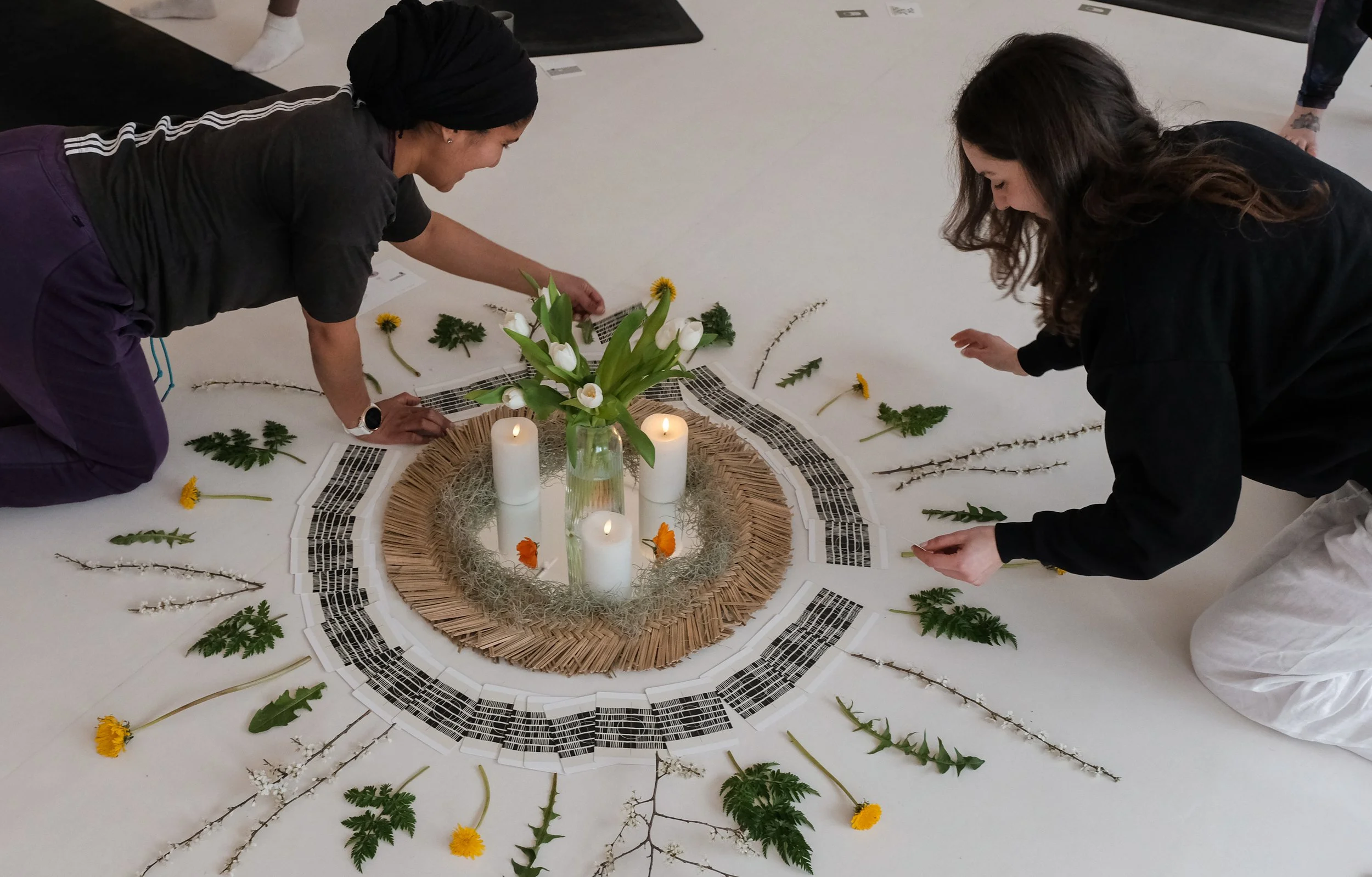 Two women kneeling on the floor arranging flowers and greenery around a circular centerpiece with candles, in a floral or art installation setting.