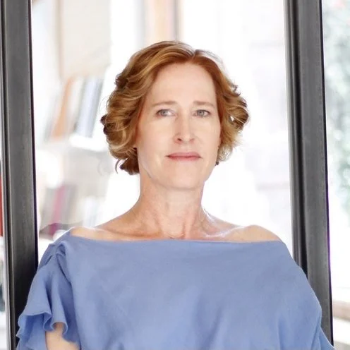 A woman with short, curly red hair looking out of a window with a thoughtful expression