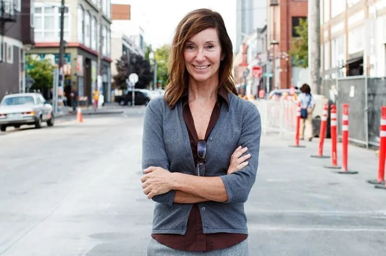 Smiling woman with brown hair standing on a city street with crossed arms, casual attire, and sunglasses hanging from her shirt.