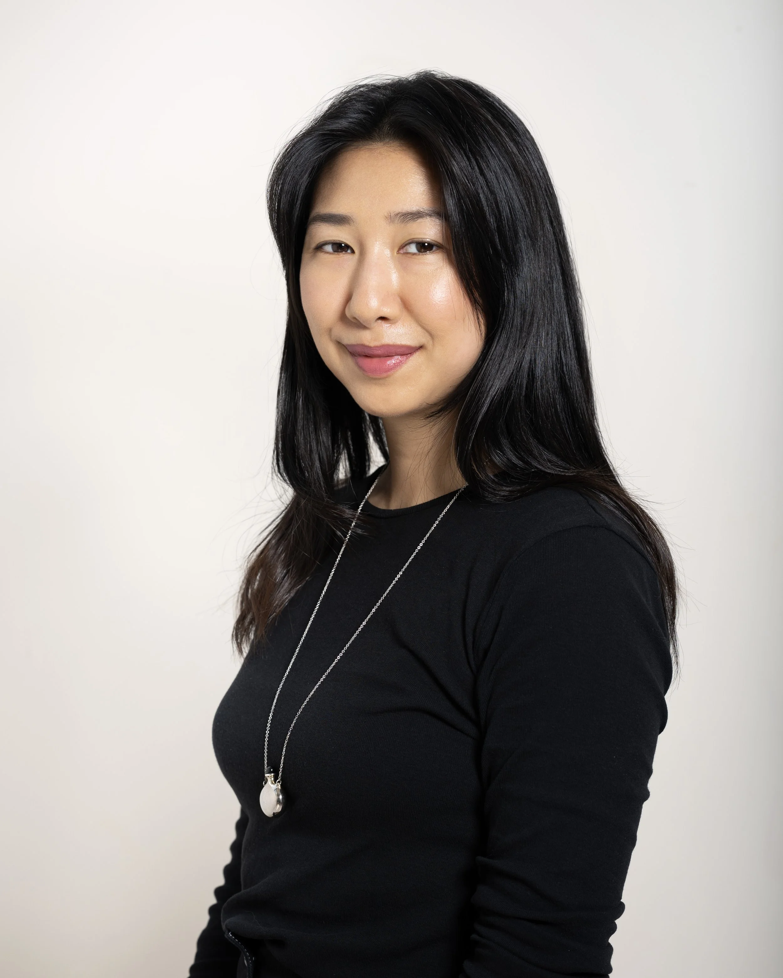A woman with long black hair wearing a black top and a silver necklace, smiling softly, against a plain white background.