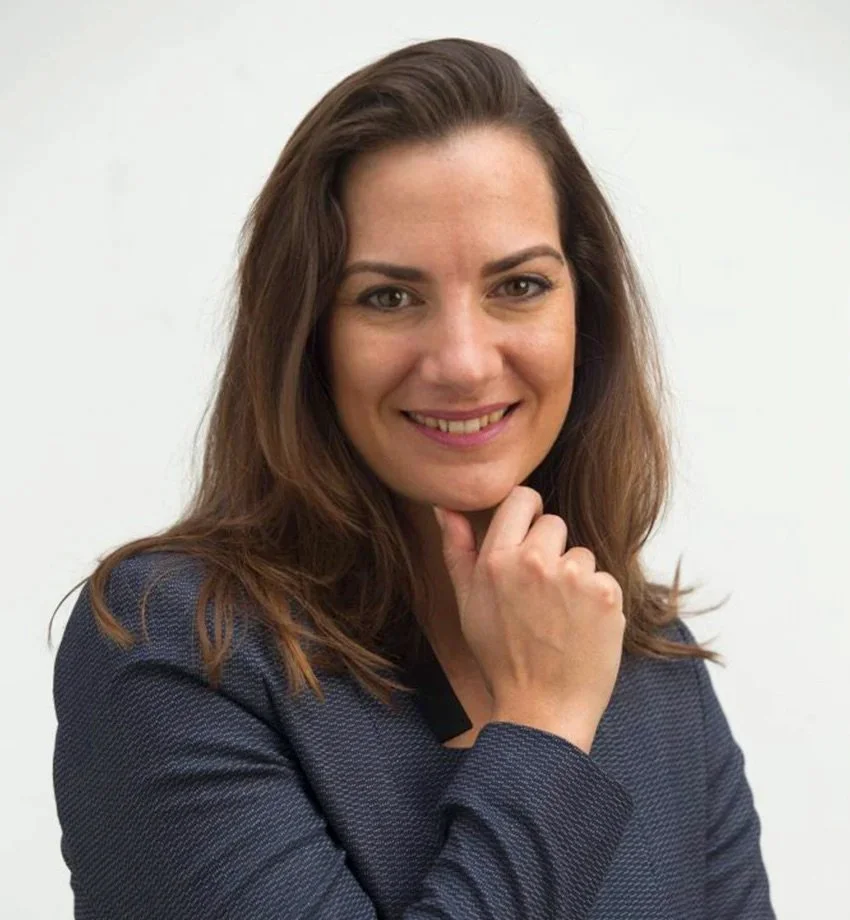 A woman with brown hair smiling, wearing a dark blazer, against a plain white background.