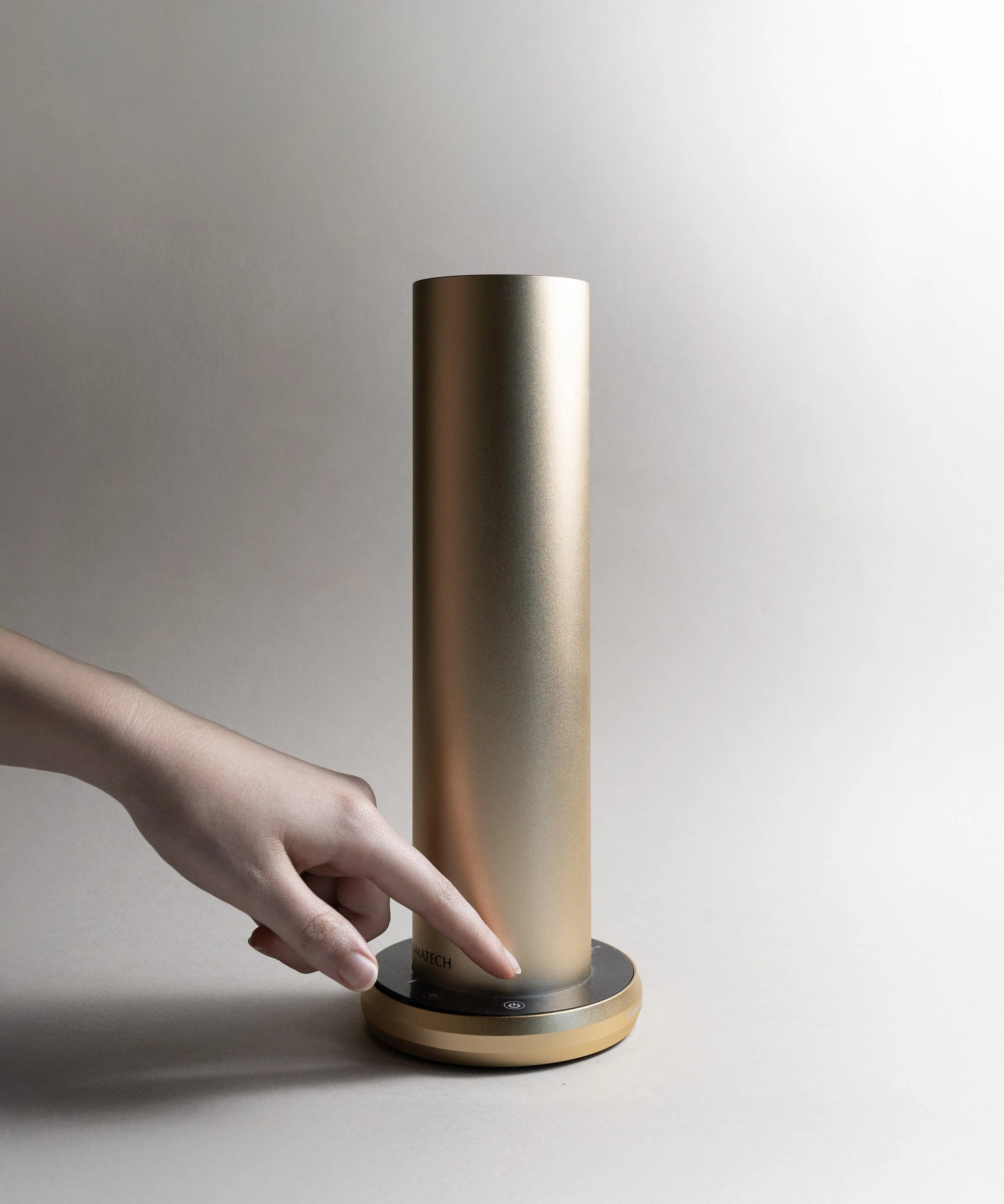 A person's hand is operating a gold-colored, cylindrical, modern-looking device with touch controls on a round base, placed on a plain neutral surface.