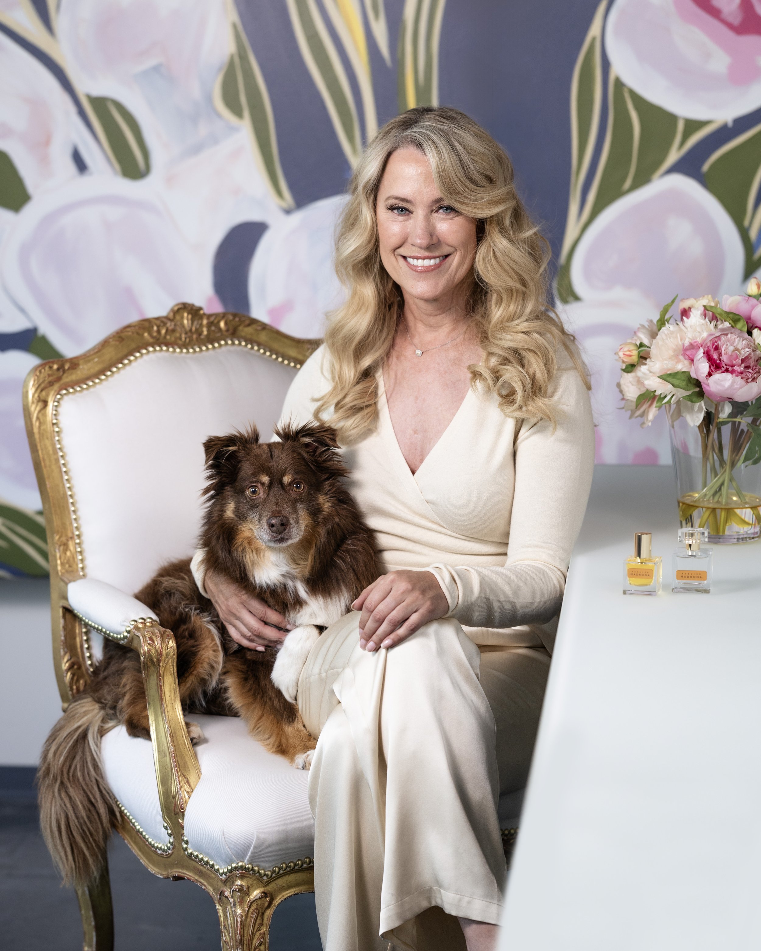 A woman with long blonde curly hair sitting on an ornate white and gold armchair, holding a brown and white dog on her lap, smiling at the camera. There is a white table with perfume bottles and a pink flower arrangement beside her, with a colorful floral mural in the background.