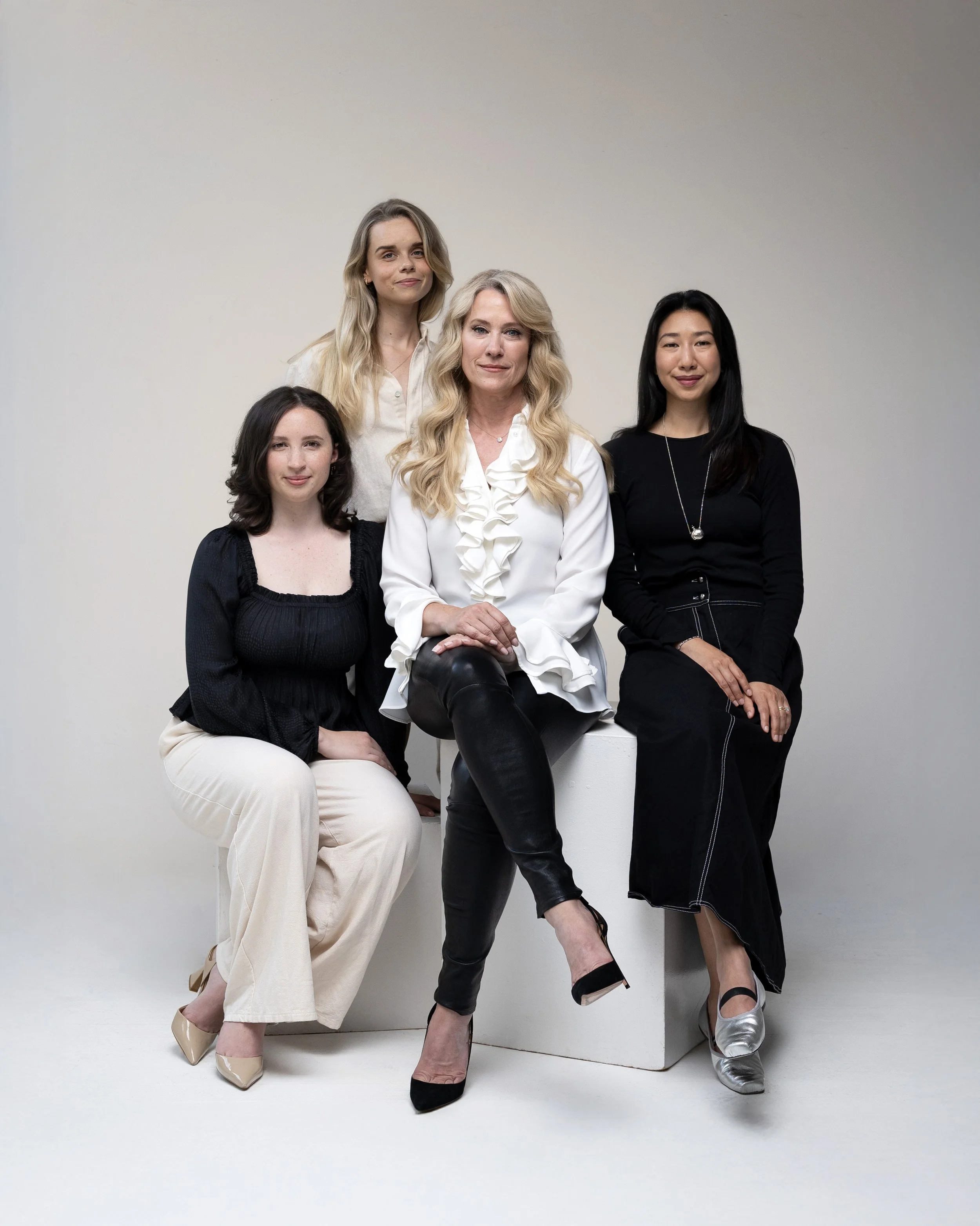 Group portrait of five women, one sitting, three standing, and one sitting on a white block, against a plain light-colored wall.