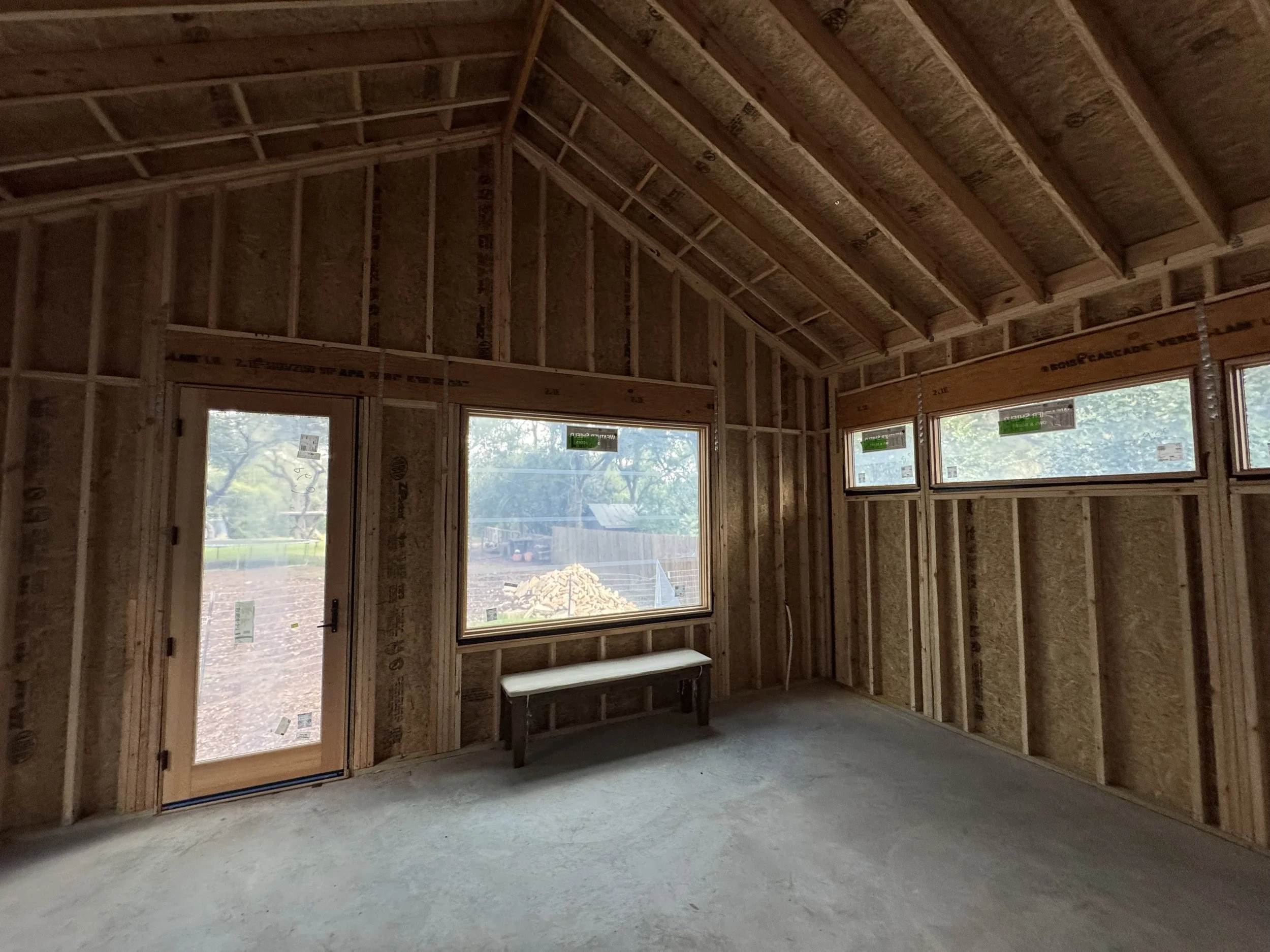Interior view of a south Austin house under construction with exposed wooden framing, large window openings showing outdoor scenery, and construction materials on the ground.