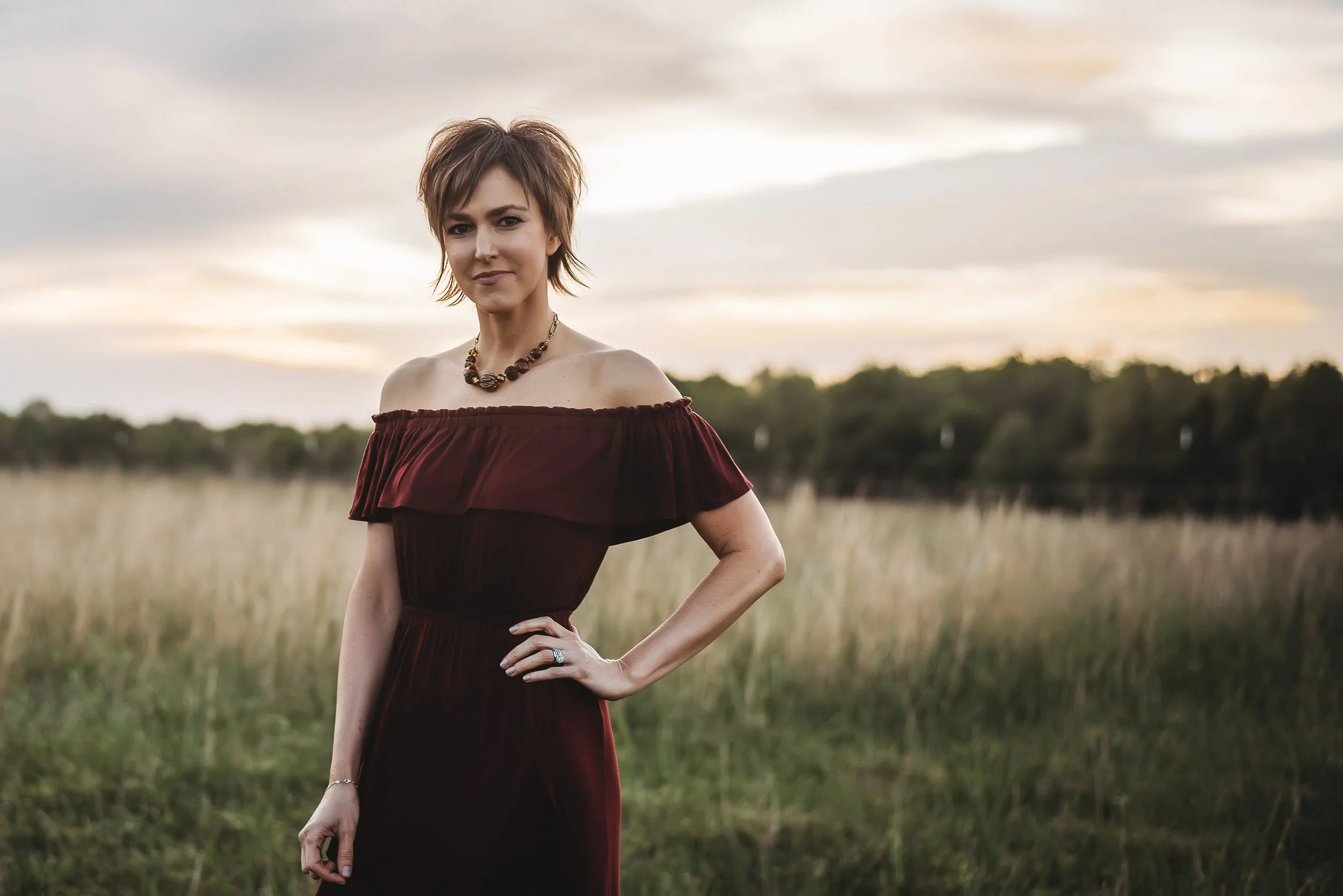 Woman in off-shoulder burgundy dress standing in a field at sunset, wearing a chunky necklace and bracelet, with short styled hair.