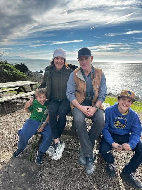 Family of four sitting on a bench outdoors near the ocean, with a cloudy sky and grassy area in the background. Two children are sitting on either side of a man and woman.