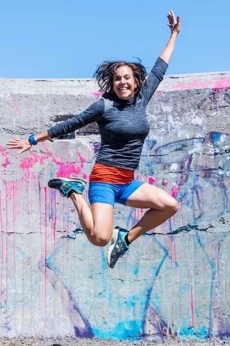 Woman jumping in mid-air with arms raised, wearing athletic clothes, in front of a graffiti-covered wall under a clear blue sky.