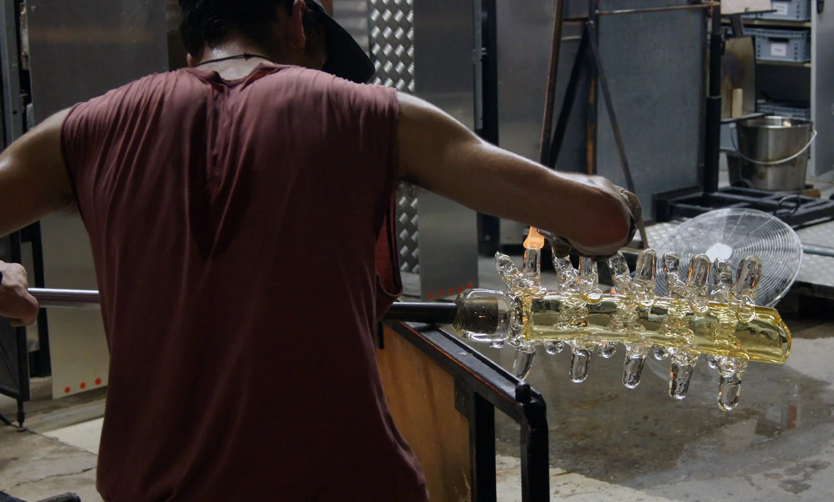 A glassblower shaping a long glass piece in a glassblowing studio.