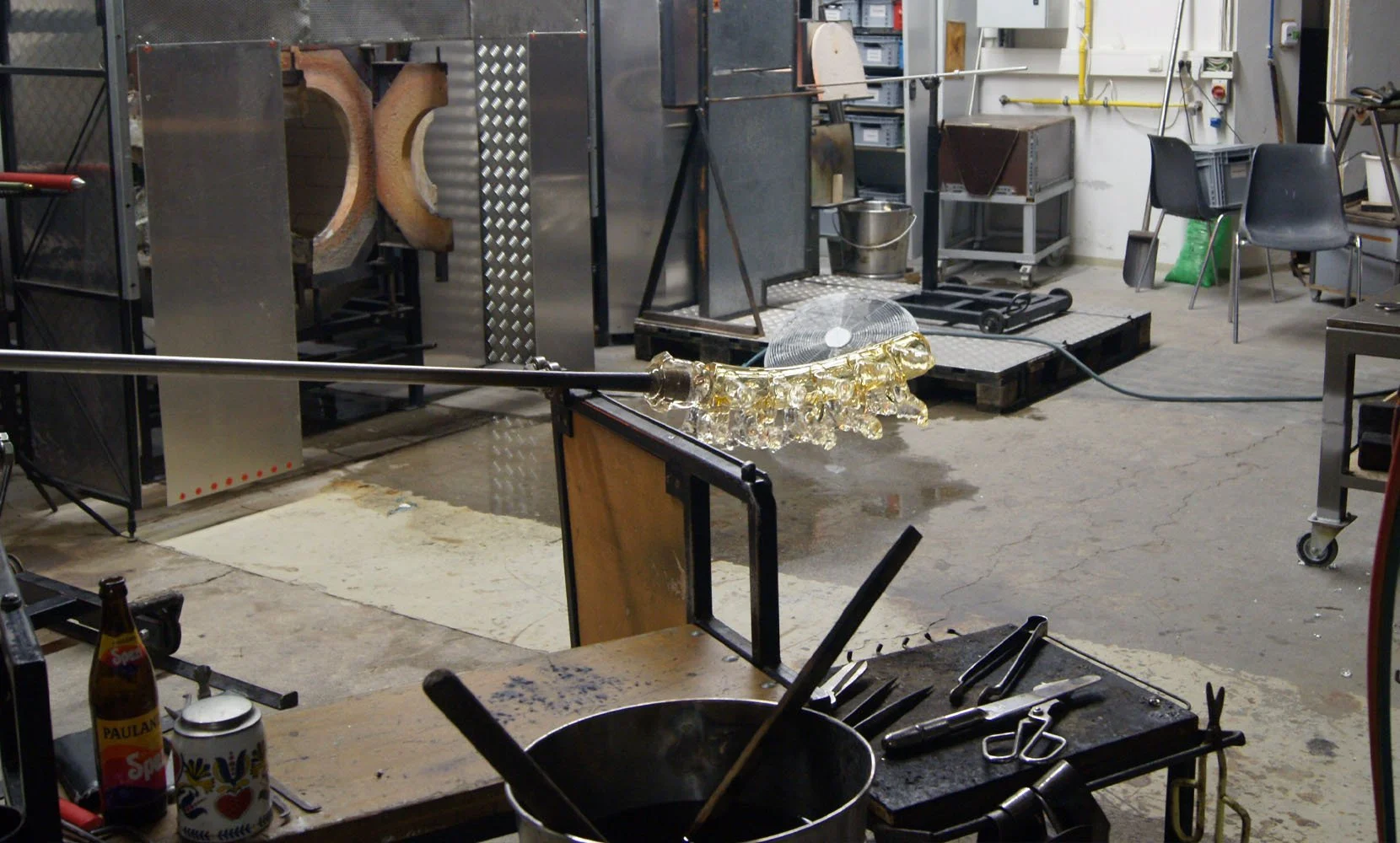An indoor glassblowing workshop with glass being shaped on a long metal blowpipe. Various tools are on a workbench, and equipment such as chairs, a fan, and shelves are visible in the background.