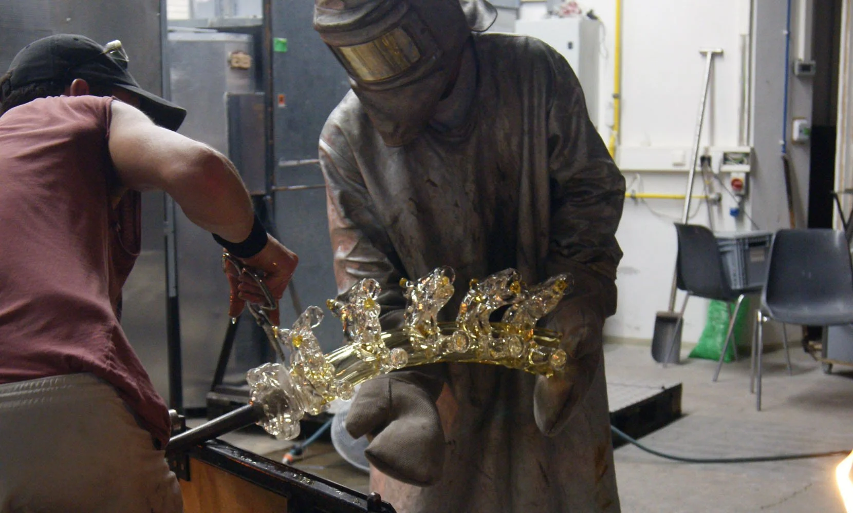 Two glassblowers working together to shape a glass sculpture in a workshop, one wearing protective gear.